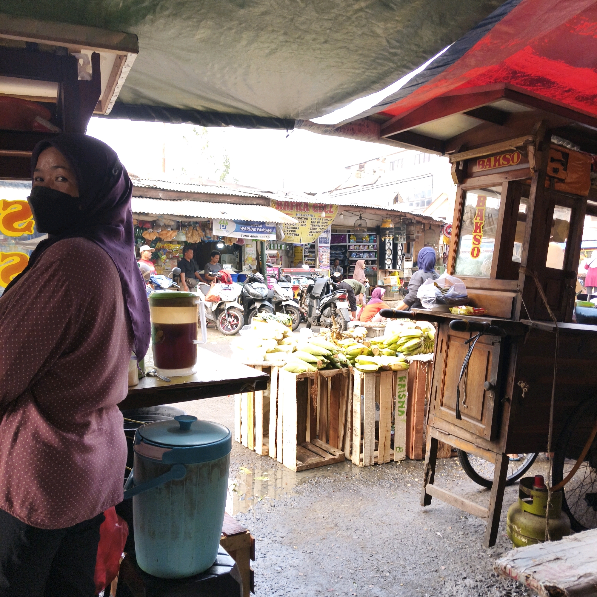 A street vendor, a woman wearing a purple hijab and a black face mask, stands at her stall in South Jakarta, Indonesia. She is positioned to the left of the frame, her body turned away from the viewer but her face looking towards the camera. In front of her is a large, blue insulated container. To the right of the woman's stall is a wooden cart selling "Bakso" (Indonesian meatball soup). The cart has a prominent sign with the word "BAKSO" in red lettering. In front of the Bakso cart are several wooden crates filled with fresh produce, including corn and bananas. Several motorcycles are parked behind the crates. In the background, other stalls and shops are visible, suggesting a busy market environment. The ground is wet and appears to be paved with asphalt. A green gas tank is visible near the base of the Bakso cart. The overall scene depicts a typical street market setting in South Jakarta.