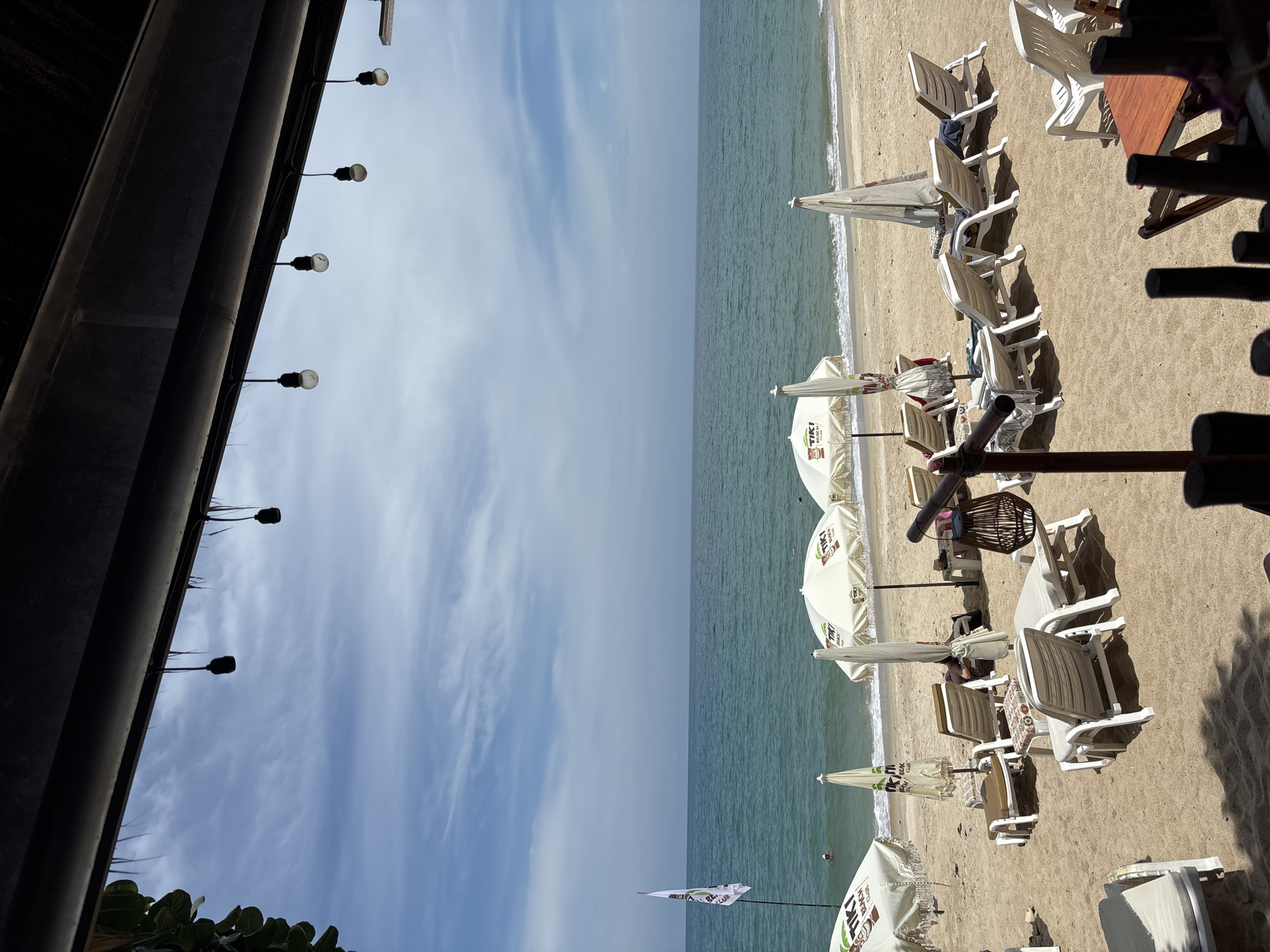 The image shows a beach scene in Baan Hua Thanon, Thailand, from a slightly elevated perspective. The left side of the image features a dark structure with a row of light bulbs, partially obscuring the sky. The right side depicts a sandy beach with several white lounge chairs and umbrellas. The ocean is visible between the beach and the sky. There are some people on the beach and in the water.