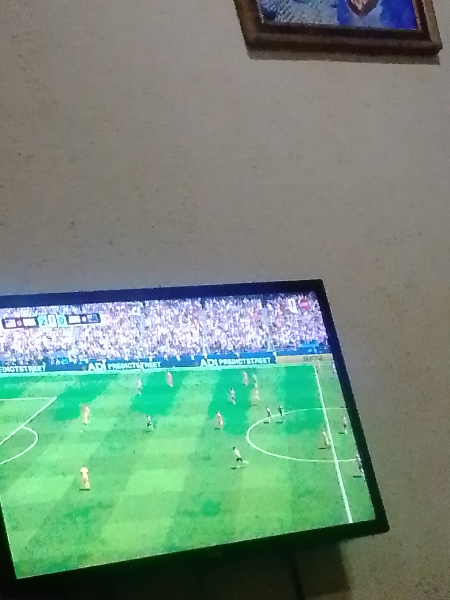 An aerial shot shows a soccer match being played on a brightly lit green field in a stadium filled with spectators. The scoreboard visible at the top of the screen shows a score of 2-0. Advertisements for "PredictStreet" are visible along the bottom of the field. The image is captured from an angle, suggesting the viewer is looking at a television screen displaying the match. The setting appears to be indoors, as indicated by the illuminated screen and the overall lighting.