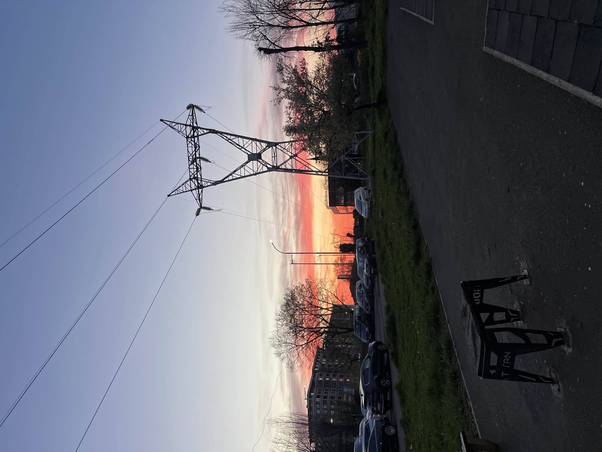 The image shows an urban scene at dusk. An electricity pylon dominates the left side of the frame, with wires stretching across the sky. The sky is streaked with colors ranging from pink and red near the horizon to blue higher up. To the right of the pylon are trees and a row of parked cars along a street. A lamppost is visible among the trees. In the foreground, on the right, there is a dark-colored bench or bike rack with the word "TITAN" visible on it. The image appears to be taken in a residential area, likely in Burgas, Bulgaria. There are no people visible in the frame.
