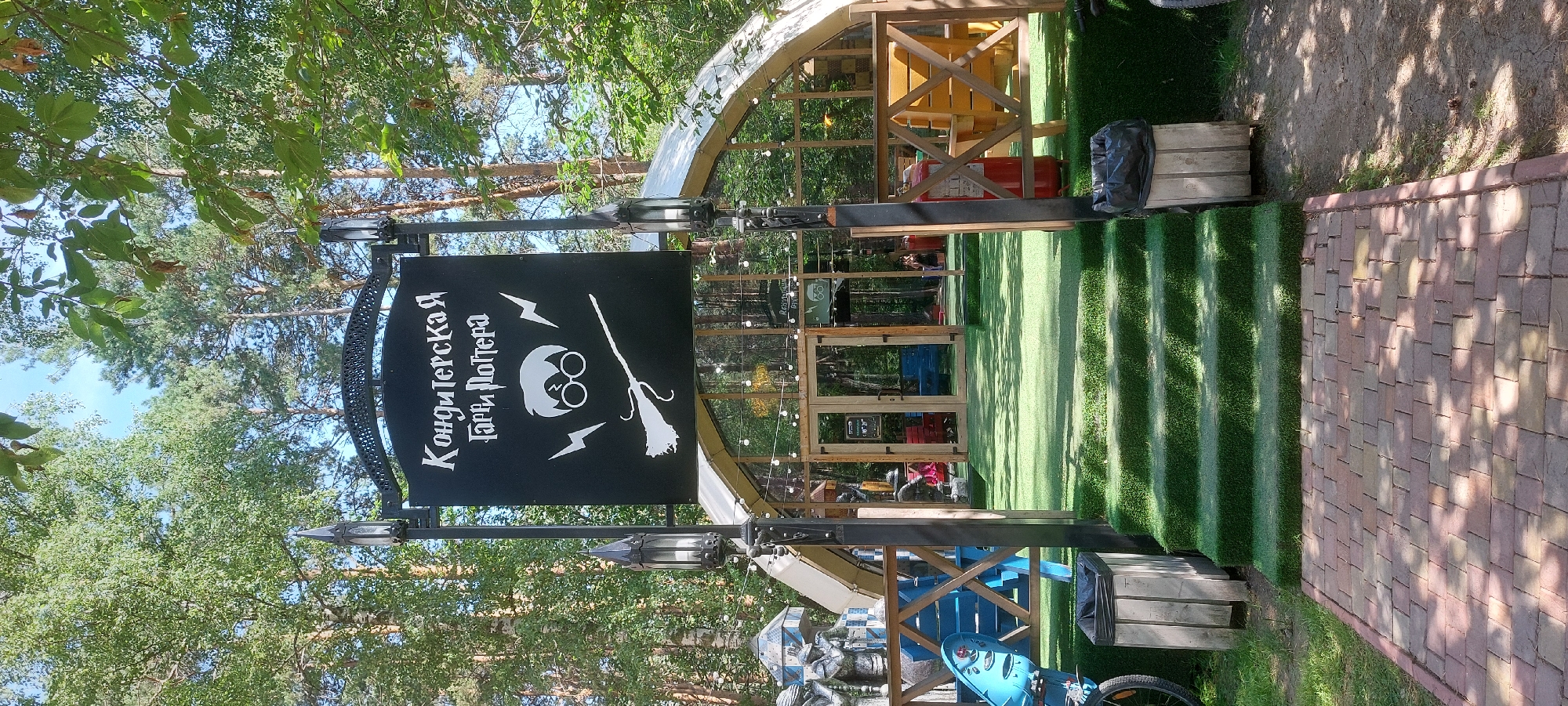 The image shows the entrance to a themed cafe or shop called "Кондитерская Гарри Поттера" (Harry Potter Confectionery) in Berdsk, Russia. The entrance features a large black sign with white text and graphics depicting Harry Potter's glasses, a broomstick, and lightning bolts. The building itself has a distinctive arched roof made of glass and wood, with string lights illuminating the exterior. In front of the building, there are steps covered in artificial turf leading up to a wooden deck with seating. The surrounding environment is a wooded area with tall trees, suggesting the location is likely in a park or recreational area within Berdsk. There are several people visible inside and near the entrance, but their actions are not clearly discernible. Various objects like trash cans, a red fire extinguisher, and parts of a playground are also present in the scene.