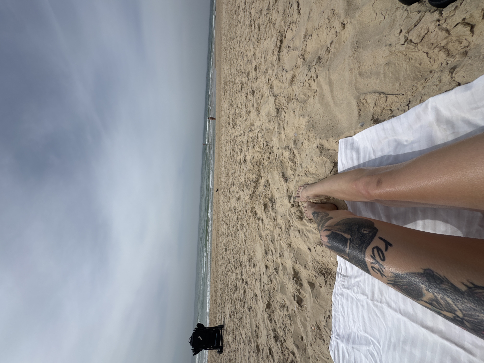 A beach scene from Bat Yam, Israel. The image appears to have been taken by someone lying on a white towel on the sand. The legs of the photographer are visible, with a large tattoo on one leg. In the distance, the sea meets the sky under a cloudy sky. The beach is sandy with small undulations. A black object, possibly a bag or piece of equipment, is visible near the shoreline. A lone figure can be seen in the distance, walking along the water's edge.