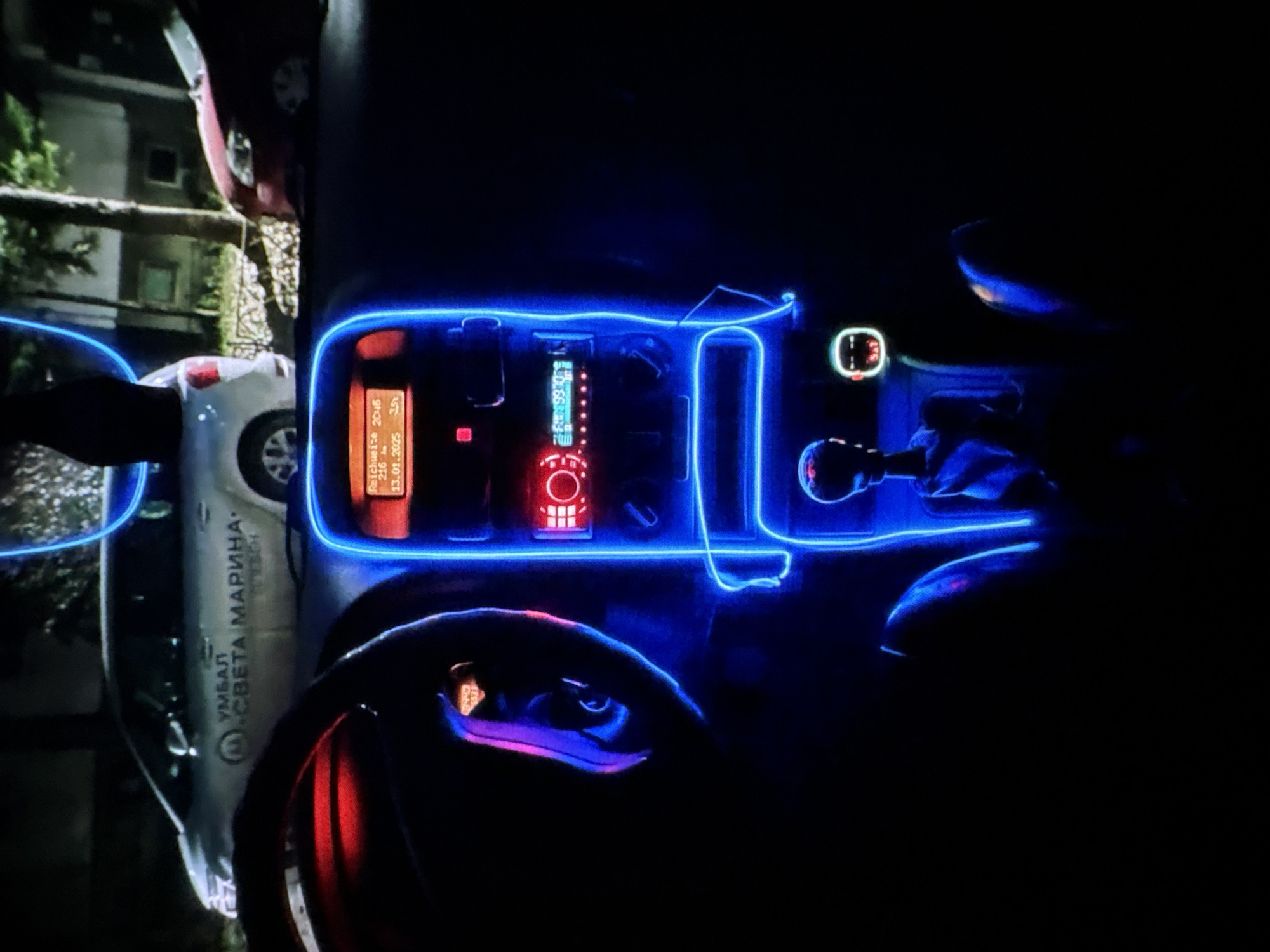 The image depicts the interior of a car at night, illuminated by blue neon lighting. The blue light outlines various components of the car's dashboard and center console. In the foreground, a person's hand is visible, interacting with a gear shifter that is also lit with purple and blue light. The dashboard features several electronic devices with displays, including a red-lit radio or media player and another screen showing text in what appears to be a Cyrillic alphabet. Part of the steering wheel is also visible, glowing with red and purple light.
Outside the car, through the windows, a building and trees can be seen, suggesting an urban or suburban setting. One of the visible text elements on the side of the car, likely a vehicle marking, reads "УМБАЛ СВЕТА МАРИНА". The overall mood is atmospheric and somewhat futuristic due to the extensive use of colored lighting. The time of day is clearly night, and there are no indications of weather conditions. The scene appears to be located in Plovdiv, Bulgaria, based on the Cyrillic text.