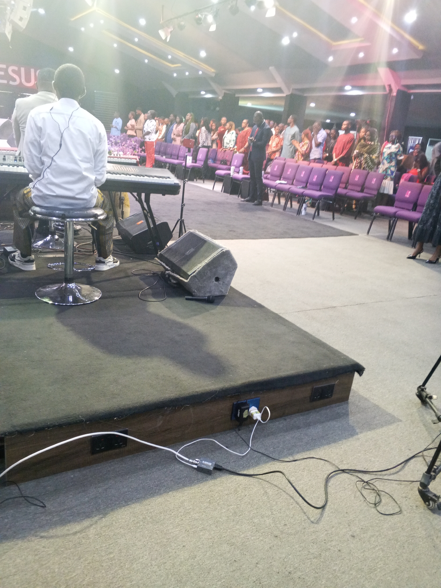 **Scene:** Interior of a large church.
**Location:** Possibly Abuja, Nigeria (city mentioned).
**People:** Group of people standing and facing forward in front of rows of purple chairs, appearing to be worshiping or listening to someone. A person in a white shirt is seated on a stool, playing a keyboard, on stage.
**Objects:** A keyboard on a stand, stage monitors, microphone on the stage floor, audio mixer on the floor, stage platform with electrical outlets and cables plugged in, purple chairs in rows, camera tripod.
**Actions:** People standing, some with hands raised. Person seated and playing keyboard.
**Interactions:** Musician playing music, congregation engaged in worship/listening.
**Environment:** Interior space with lighting fixtures and high ceiling. The space appears to be a church or religious assembly hall.