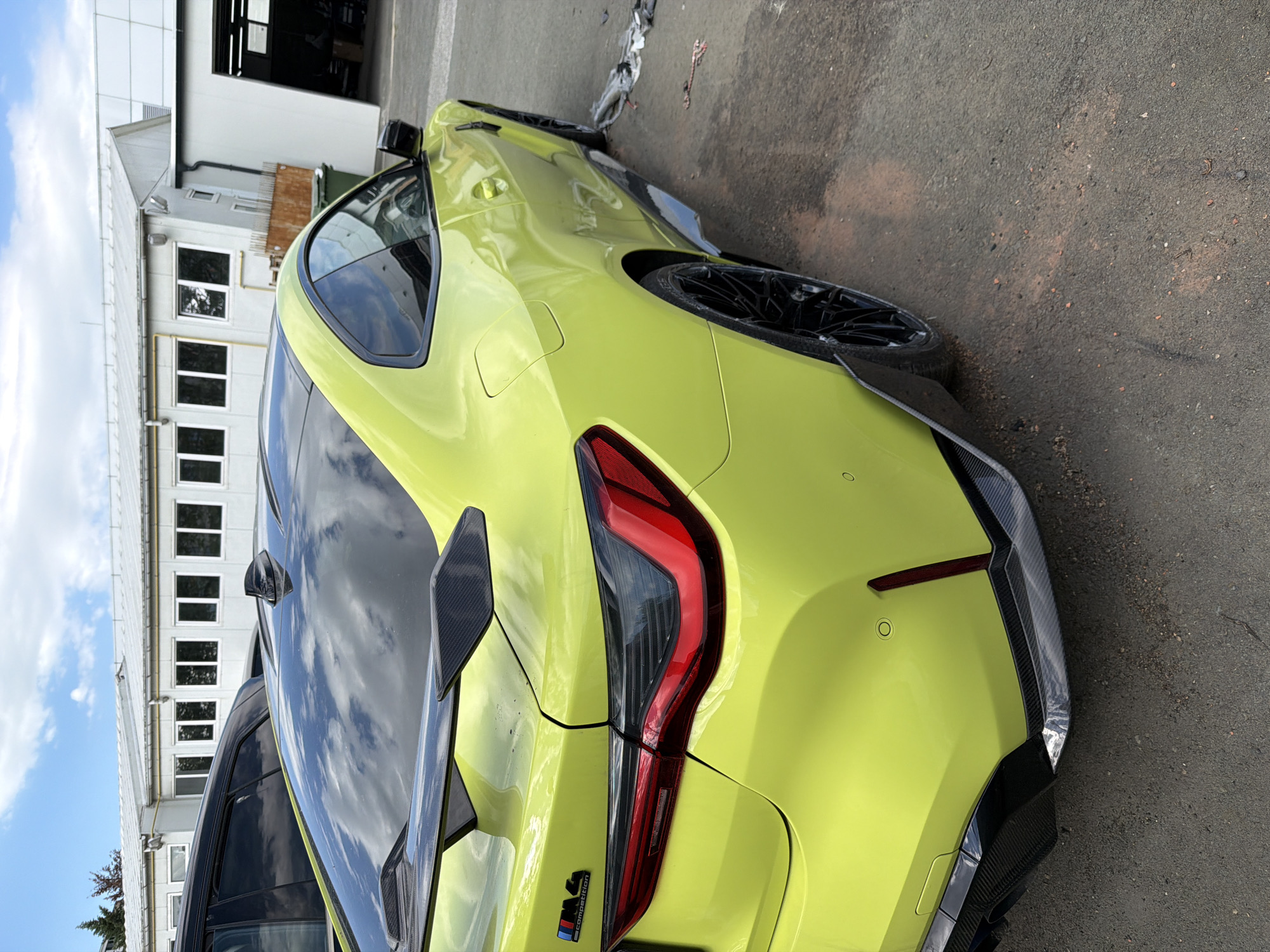 A yellow BMW M4 Competition coupe is parked outside a building. The building has white walls with regularly spaced rows of windows. The car is parked on an asphalt surface. There are no people visible in the frame. Based on the "M4 Competition" badge, the car is likely a high-performance variant. The image does not provide enough information to confirm the location in Burgas, Bulgaria, with certainty.