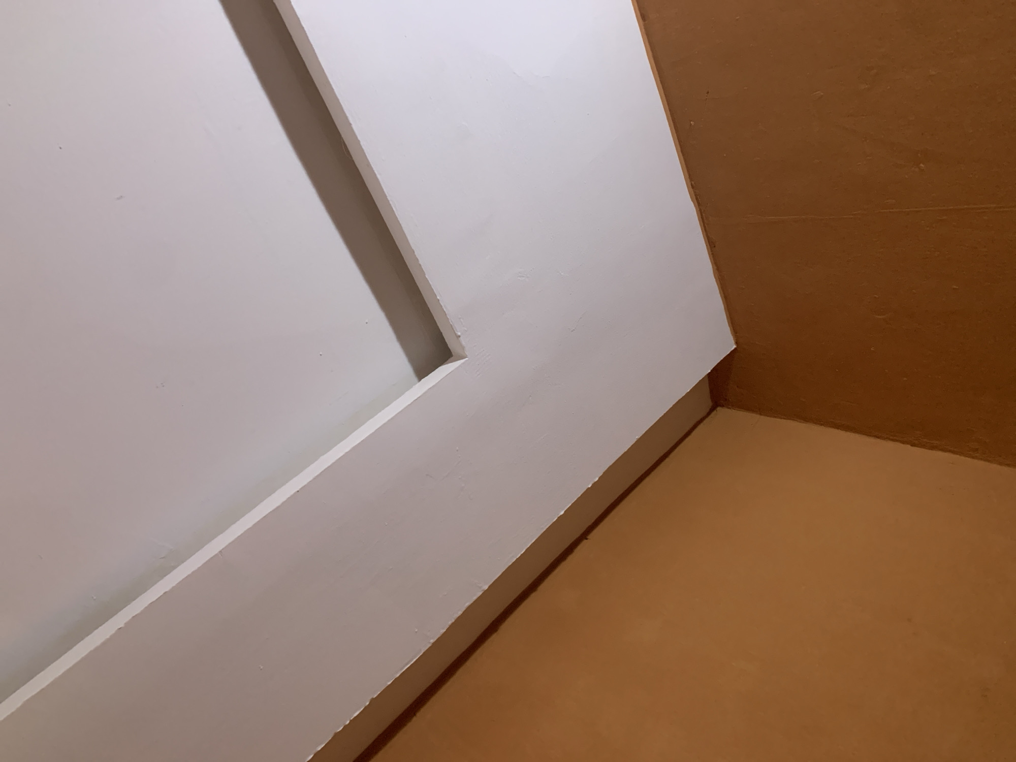 The image shows an interior view of a corner where two walls meet the ceiling. The walls are painted a warm, light brown or orange color, and appear to have a textured finish. The ceiling is white and features a recessed element with a shadow, suggesting a layered or tray ceiling design. There is a white trim or molding that runs along the intersection of the walls and the ceiling. The lighting appears to be from above, casting subtle shadows. There are no people or any other discernable objects in the scene. The perspective is upward, looking towards the ceiling.