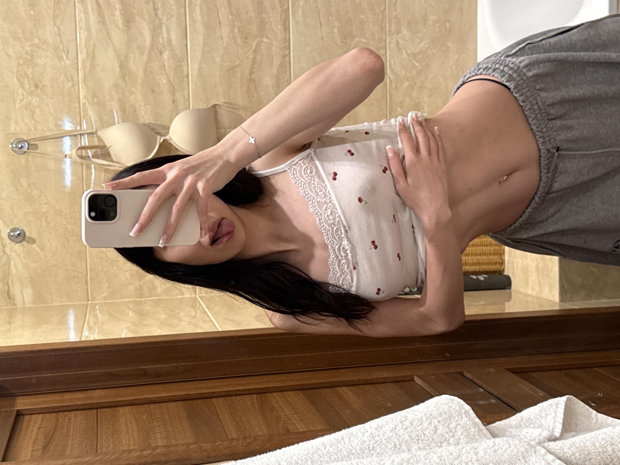 The image depicts a woman taking a mirror selfie in a bathroom. She is wearing a white tank top with a cherry print and lace trim, and grey sweatpants. Her tongue is sticking out slightly, and she is holding her stomach with one hand. A white towel hangs on the left side of the frame, and a beige bra is visible hanging on the tiled wall behind her. The overall environment appears to be a residential bathroom. There is no specific indication of location within the city of Khimki, Russia.