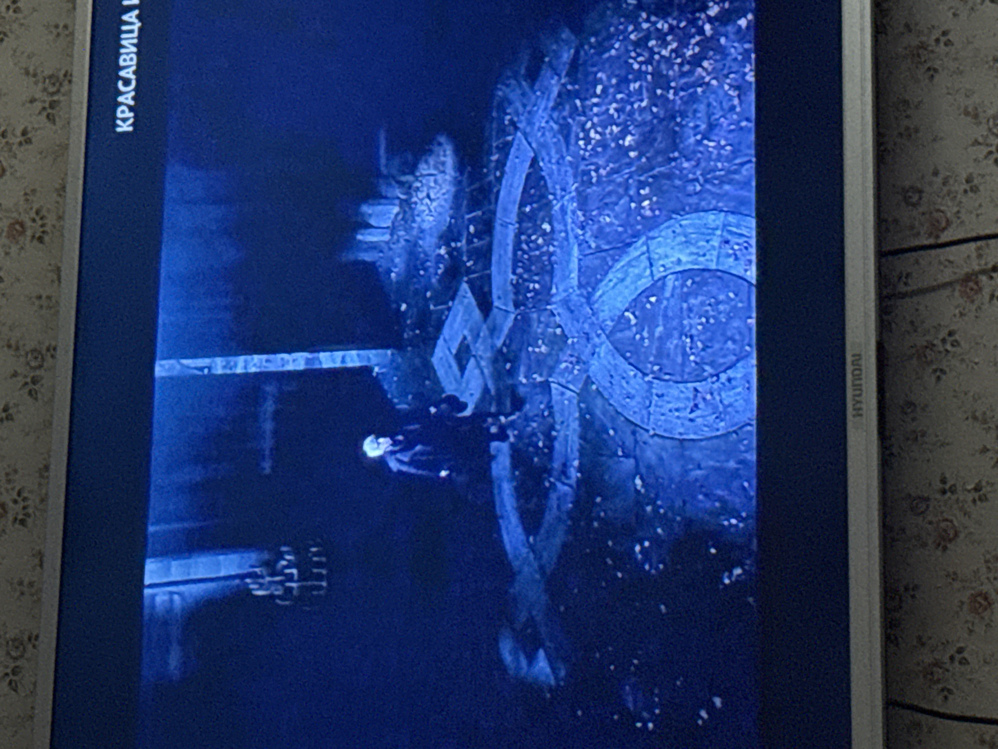 The image shows a television screen displaying a scene from what appears to be a movie or television show. The scene is dark and illuminated by blue light. A figure, possibly a person, stands on a stone structure featuring a prominent circular or repeating pattern. The structure appears to be situated in an outdoor environment that is either covered in or adjacent to trees. "Красавица И" is visible on the left edge of the screen. The Hyundai logo can be seen on the lower right edge. The location is potentially Mytishchi, Russia.