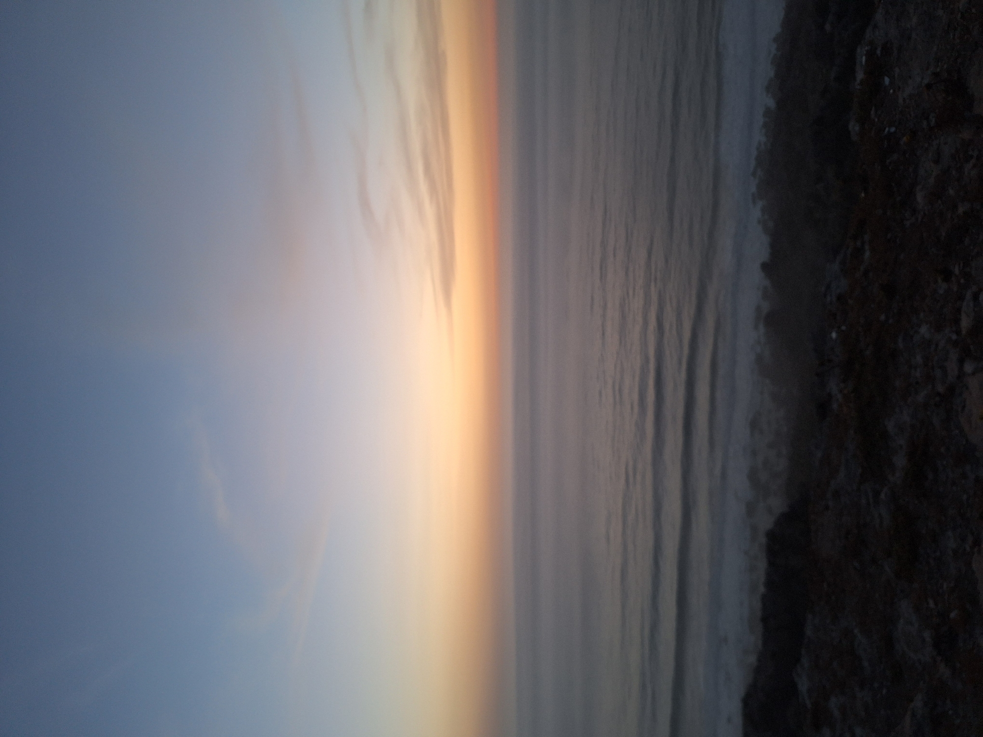 The image depicts a serene seascape at dusk, likely captured from a coastal vantage point in Ahrara, Morocco. The sky transitions from a pale blue at the top to soft hues of orange and pink near the horizon, suggesting the sun has recently set or is about to rise. A few wispy clouds are visible in the upper part of the sky. Below the sky, the ocean stretches out, appearing calm with gentle waves creating subtle ripples. The water is a muted grey-blue, reflecting the twilight colors. In the foreground, the image shows a dark, rocky terrain, characteristic of a rugged coastline. This rocky area is sparsely covered with what appear to be low-lying vegetation or scrub. The overall atmosphere is tranquil and undisturbed, with no people or man-made structures readily apparent, emphasizing the natural beauty of the coastal landscape.