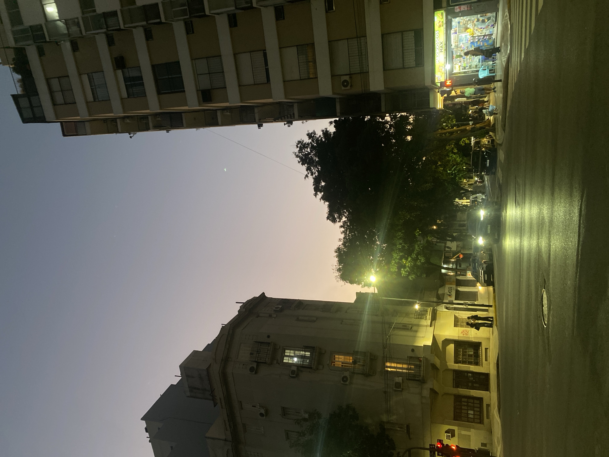 This image depicts the intersection of Av. Córdoba and Jean Jaurès in the Balvanera neighborhood of Buenos Aires, Argentina, at dusk. The sky exhibits a light purple-orange gradient near the horizon transitioning to dark blue overhead, with a faint crescent moon visible in the upper right quadrant.
To the left, a multi-story, light-colored older building with a corner bay window, balconies with dark iron railings, and numerous externally mounted air conditioning units occupies the street corner. A red traffic light is active on a pole attached to this building's facade, and several interior lights are visible through its windows.
To the right, a taller, modern multi-story building features a light beige facade with white horizontal bands and balconies. Its ground floor houses a brightly illuminated "ANTOJITOS MAX" kiosk/convenience store, displaying merchandise through its windows. A light blue chair is positioned on the sidewalk outside the kiosk.
The asphalt street is reflective, possibly wet, with a circular manhole cover in the foreground and white crosswalk markings on the right. Two vehicles with illuminated headlights are present in the mid-ground on the road. A prominent streetlight in the center casts a strong yellow-green glow over the intersection and onto the dense foliage of a large tree positioned centrally.
Approximately seven to nine individuals are visible on the sidewalks. Two individuals are standing and conversing near the older building on the left. On the right, several individuals are gathered near the "ANTOJITOS MAX" kiosk, with one person appearing to be stooping or crouching. Electrical wires traverse the upper portion of the frame.