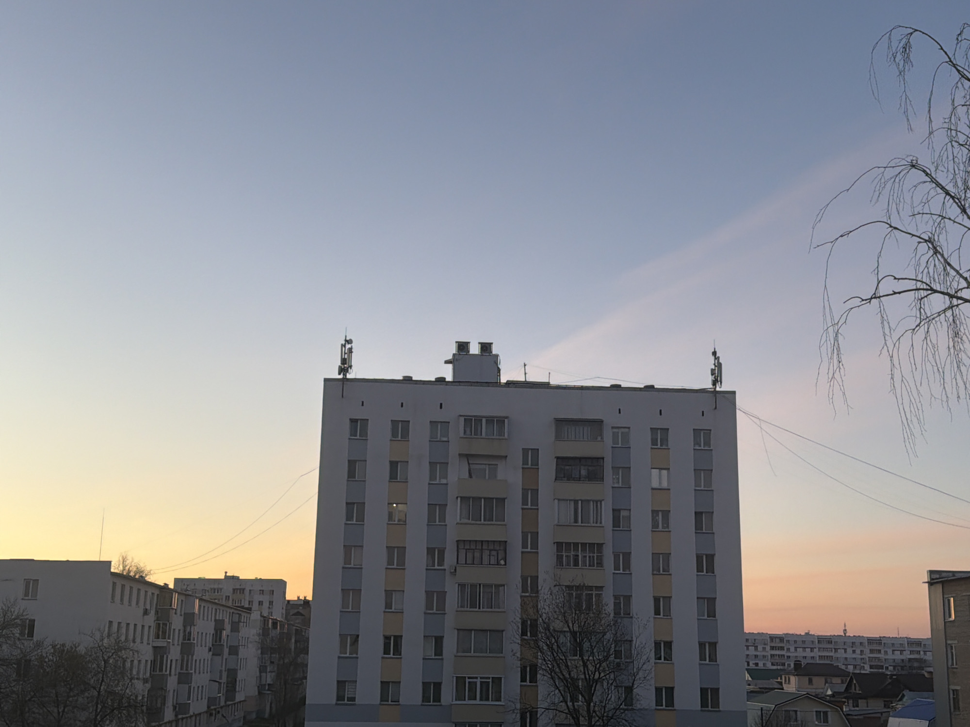 The image depicts a cityscape at dusk, likely in Naberezhnye Chelny, Russia. A tall, rectangular apartment building dominates the foreground, featuring a white facade with vertical stripes of color and rows of windows. Antennas are visible on the building's roof. A network of wires extends from the top of the building. Other similar residential buildings appear in the background. The sky transitions from a pale blue to a warm orange near the horizon. Bare tree branches are visible in the foreground on the right side.