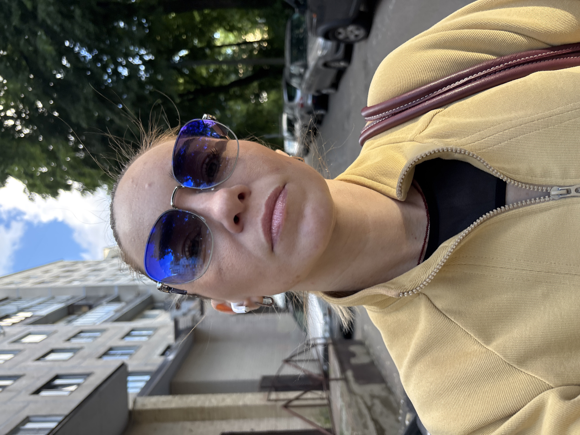 A woman wearing sunglasses and a yellow jacket is walking on a street in an urban environment. She has earbuds in both ears and is carrying a bag over her shoulder. The background shows a building, trees, and parked cars. The image appears to be a selfie taken at eye level.