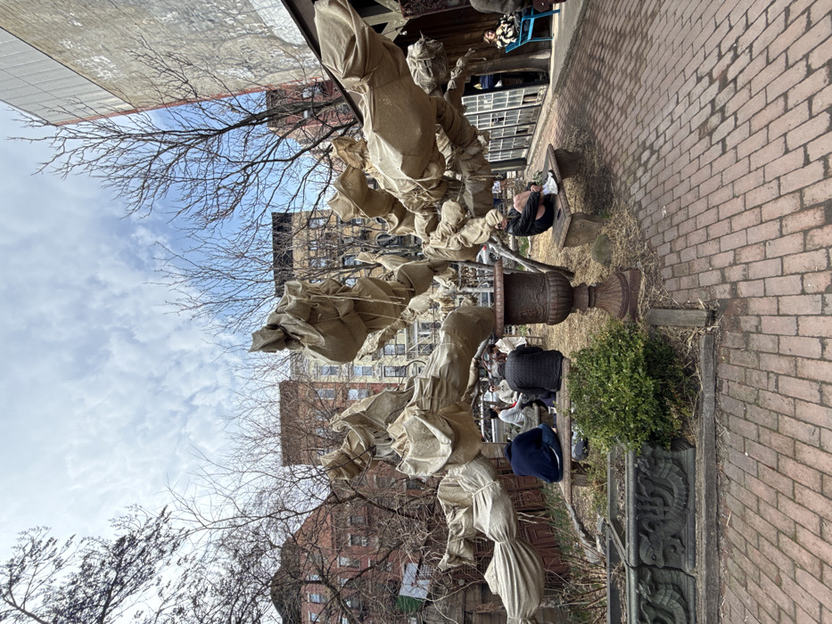 This image depicts an outdoor urban garden, identified as the Elizabeth Street Garden, located in Nolita, Manhattan, New York, United States of America. The scene is captured on a partly cloudy day, likely during late winter or early spring, as most background trees are bare.
In the center of the frame, a prominent deciduous tree is entirely wrapped in light-brown coarse fabric, resembling burlap, which is secured with twine, giving it a distinct sculptural appearance. Surrounding this wrapped tree are several rustic grey stone benches. Eight individuals are present in the garden. Five are seated on these stone benches: four are clustered on a bench to the left of the tree, engaged in conversation, one wearing a dark blue hoodie and another a dark patterned jacket. A fifth individual is seated alone on a bench to the right, facing left, dressed in dark shorts and a black jacket, appearing to adjust their white sneakers. Two more individuals are partially visible further to the right near a building facade, one seated on a blue chair.
In the foreground, a large, ornate, dark-colored cast-iron urn on a pedestal stands prominently. To the left foreground, a low, rectangular stone planter, adorned with carved mythological or animal figures, holds a small, vibrant green bush. The ground is paved with red brick laid in a herringbone pattern, transitioning to bare earth and dry vegetation in the immediate vicinity of the benches and the tree.
The background features a row of multi-story residential buildings, primarily brick and brownstone, showcasing various architectural styles and colors under a bright, partly cloudy sky. Bare tree branches are silhouetted against the sky and the buildings. To the far right, a taller, light-colored building with multiple large, vertical windows is visible. The overall scene suggests a tranquil, communal urban green space.