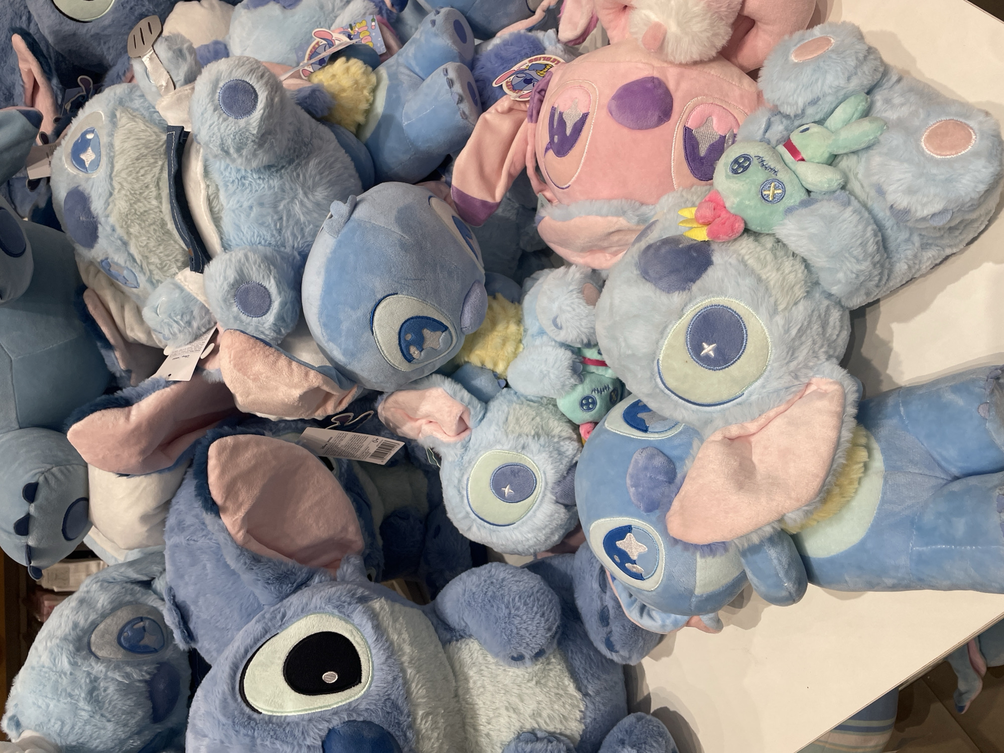 A collection of blue and pink plush toys resembling the character Stitch from Disney's Lilo & Stitch are piled on a white surface, possibly a retail display. The toys are of various sizes, with some being large and others smaller. They are predominantly blue, with some pink variants also visible, particularly one larger pink Stitch-like toy in the foreground. Several of the toys have large, expressive eyes with distinctive Stitch markings. Some of the plushies are adorned with small accessories, such as a tiny green voodoo doll-like figure holding a pink and yellow accessory, perched on the chest of a light blue plush. The background is blurred, suggesting a store environment. The lighting is bright and even, indicating an indoor setting. This scene is likely from a store in Davao City, Philippines, given the context provided.