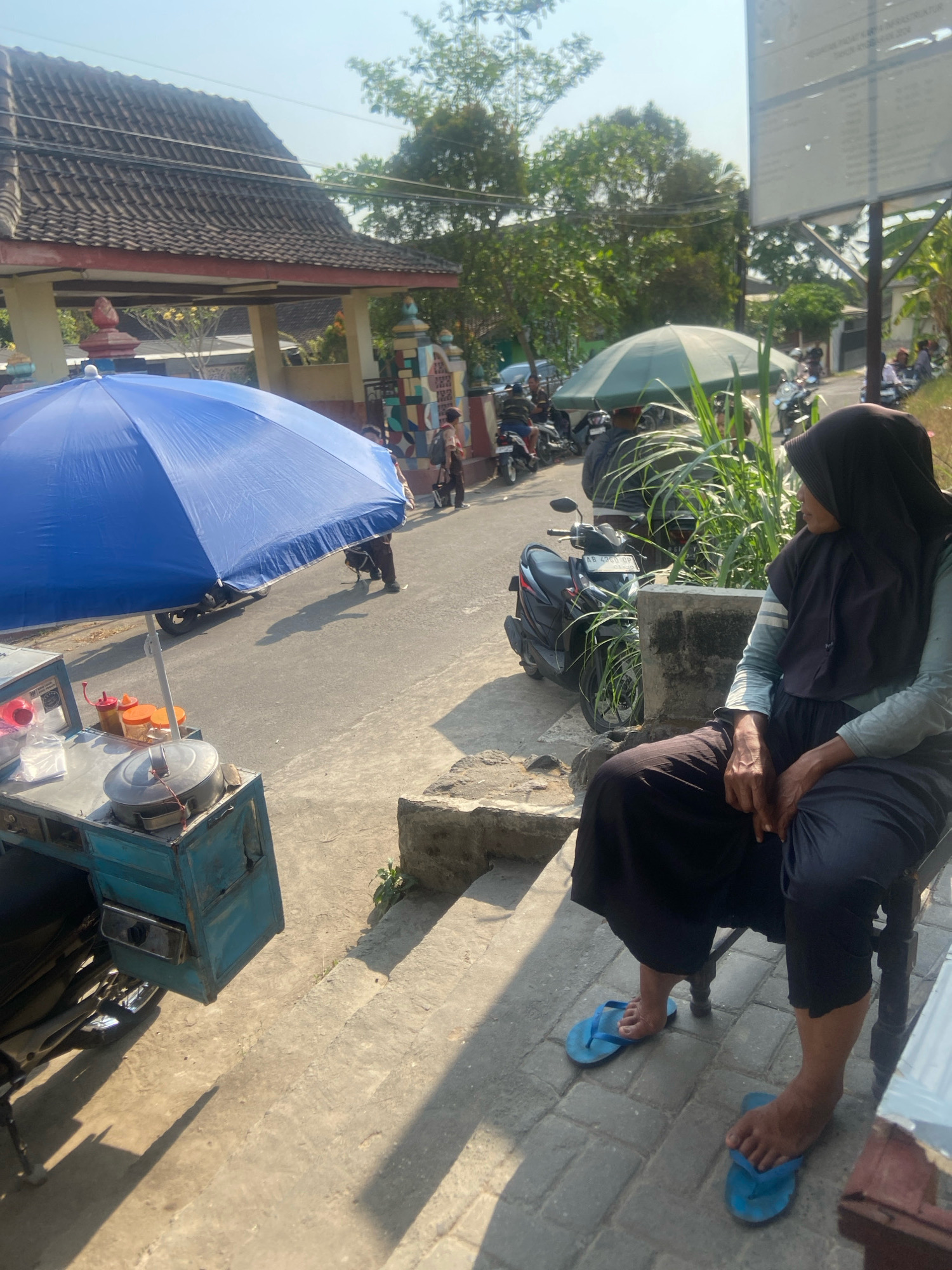 This image depicts a street scene in Sleman, Indonesia, under a clear, sunny sky. In the foreground, a woman wearing a dark hijab and dark clothing, including blue flip-flops, is seated on a chair. To her left, a blue umbrella shades a food cart with an attached motorcycle. The cart holds containers of food items and a large metal pot.
Behind the food cart, a paved road is visible, with several motorcycles parked on the side. Further back, a building with a tiled roof and pillars stands, and beyond that, trees and more buildings are present. The overall environment suggests a bustling street or market area. The woman is looking to the right, with her hands resting in her lap. The lighting indicates it is daytime, possibly late morning or early afternoon, given the shadows.