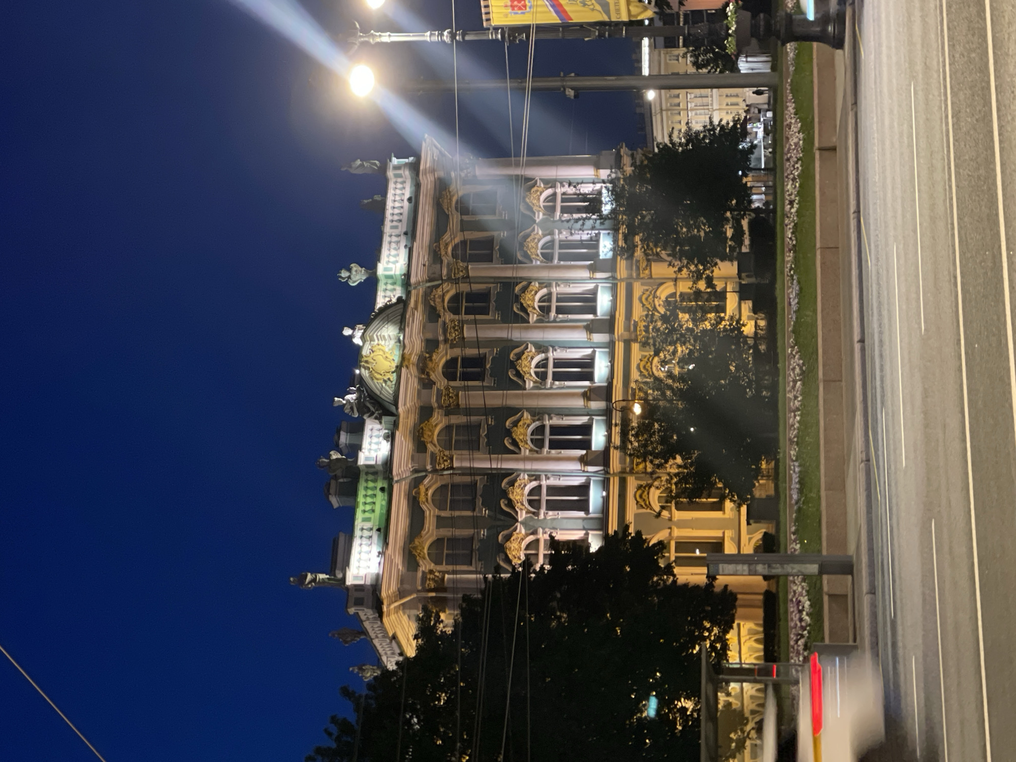 The image displays a grand urban night scene, likely in Saint Petersburg, Russia, characterized by its distinctive architecture and atmospheric lighting. Dominating the left side of the frame is a magnificent multi-story building, showcasing elaborate classical or Baroque design. Its facade, possibly light grey or green under the night lights, features numerous arched windows surrounded by rich, gilded ornamentation. Statues and sculpted figures adorn the roofline, adding to the building's regal appearance. Many windows are illuminated from within, casting a warm glow.
To the right, a wide, paved road is visible with distinct white lane markings. Separating the road is a well-maintained grassy median strip, interspersed with flowering plants and shrubs. Tall, dark street poles punctuate the scene; one prominent pole on the right side emits bright, circular lights that create noticeable light rays across the dark blue sky. Another street pole further down features a banner or flag with a crest or emblem, but any text on it is illegible due to the distance and resolution. Dark trees are scattered along the median and in front of other less distinct buildings in the background.
The time of day is night, indicated by the deep blue, clear sky and the reliance on artificial illumination. The soft ambient light suggests either late twilight or potentially the "white nights" phenomenon common in Saint Petersburg during summer. The overall mood is one of majestic tranquility and historical grandeur. No people or vehicles are visible, contributing to the serene stillness of the urban landscape.