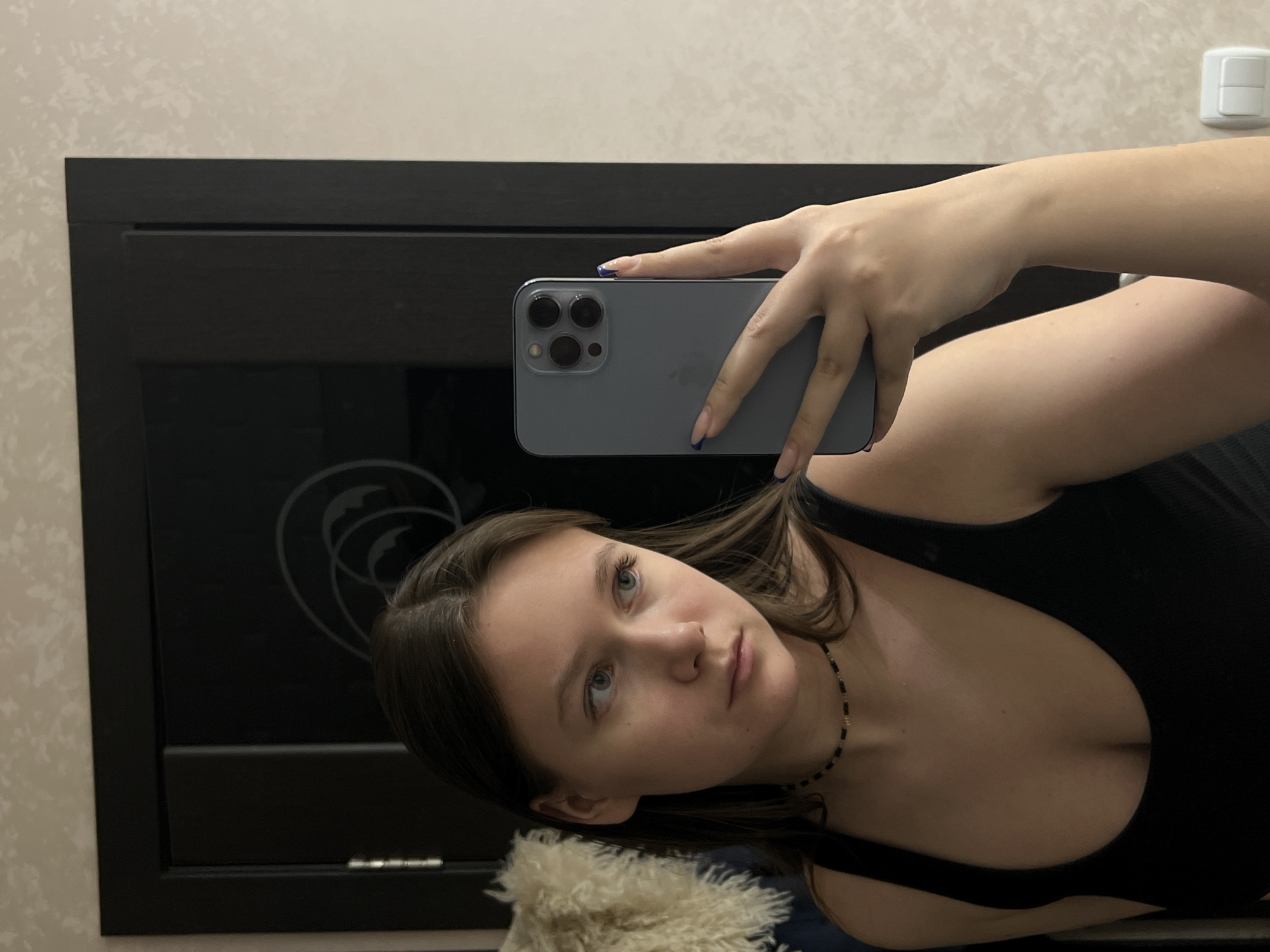 A young woman is captured in what appears to be a mirror selfie, rotated 90 degrees clockwise. She has fair skin, brown hair, and light-colored eyes, looking directly towards the camera with a neutral expression. She is wearing a black tank top with a scoop neck and a thin black choker necklace. Her fingernails have blue tips.
She is holding a grey-blue smartphone, identifiable as an iPhone with its characteristic triple camera system and the Apple logo visible on the back. Her hand is positioned to hold the phone, suggesting she is taking the photo.
The background shows an indoor setting. A large, dark, reflective surface, possibly a mirror or dark furniture, occupies a significant portion of the image. This surface reflects a partial circular design. To the left and top of this dark surface, a light-colored wall with a subtle textured wallpaper pattern is visible. In the upper right corner, a white rectangular electrical outlet or light switch is mounted on the wall. A light-colored, fluffy item, possibly a cushion or part of clothing, is partially visible in the lower-left foreground.
The lighting suggests an interior setting, possibly daylight or ambient room light. No explicit text is visible other than the Apple logo on the phone. Given the context, the location is Chaikovsky, Russia.