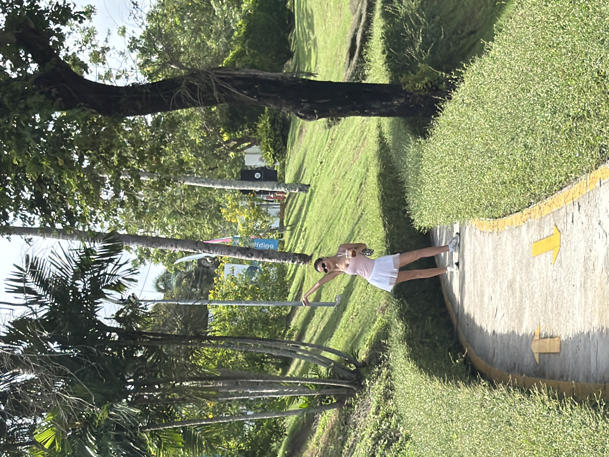 A woman stands on a paved path with directional arrows in Choeng Thale, Thailand. She wears a light pink top and white skirt. Palm trees and greenery flank the path. A large tree with a dark trunk is visible in the background. Banners or signs with lettering are visible behind her.