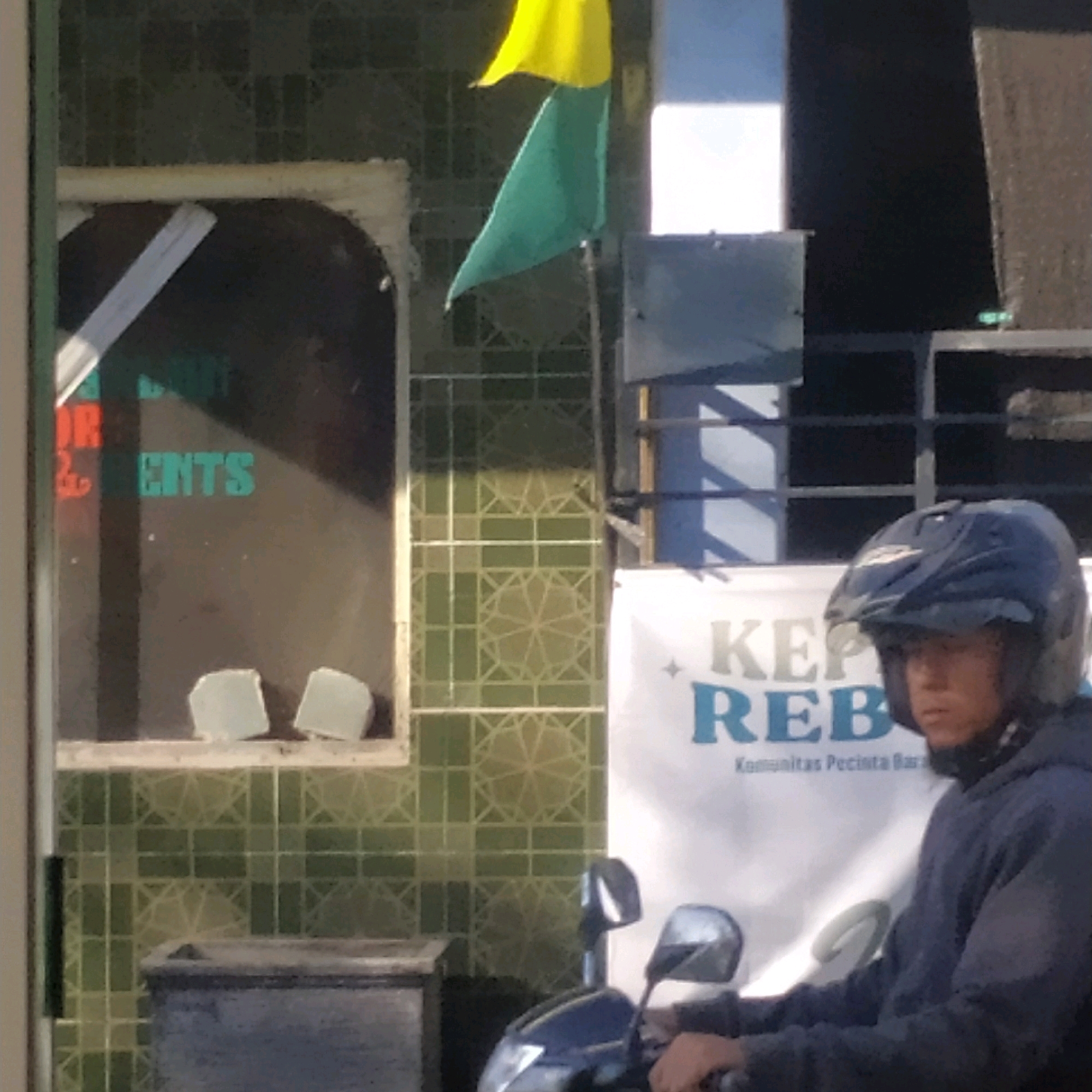 The image depicts a street scene, likely in Batan, Indonesia, where a young man wearing a blue hoodie and a black helmet is sitting on a motorcycle. He is looking to the right of the frame. The background features a building with green tiled walls and a window. A sign with red and green text, possibly indicating a business, is visible in the window. To the right of the building, a white banner with blue text, "KEP", "REB", and smaller text below, is displayed. Two flags, one yellow and one green, are flying from a pole in the upper left portion of the image. The overall lighting suggests it is daytime.