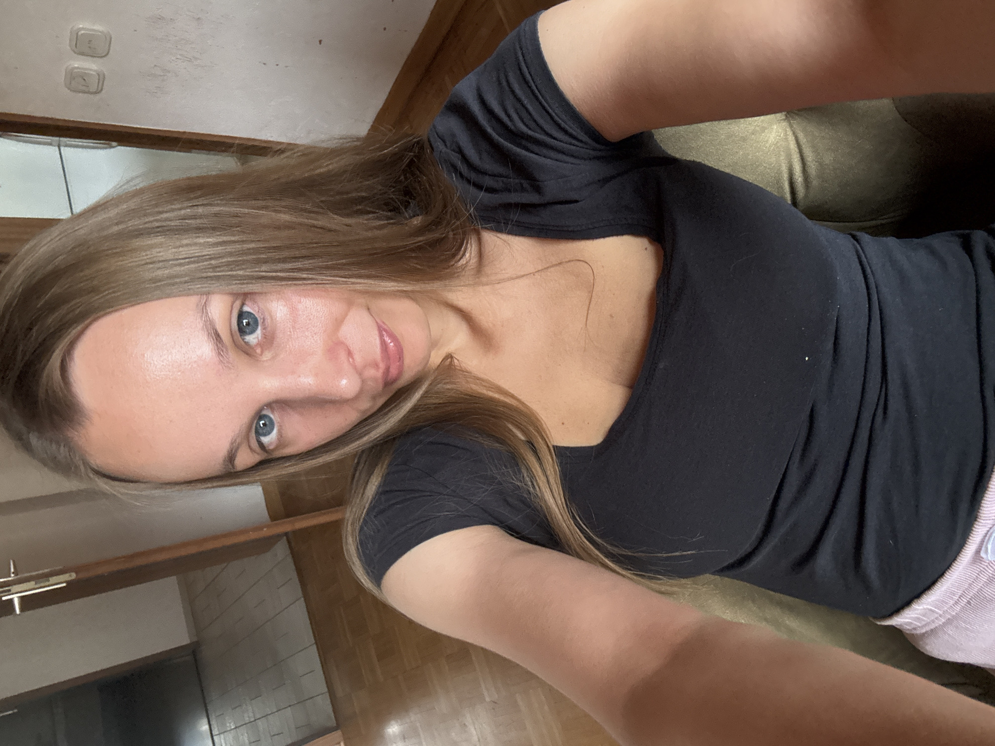 A woman with long blonde hair and blue eyes is taking a selfie indoors. She is wearing a black t-shirt and light pink shorts. She is seated in a chair with a green upholstery. The background shows a doorway leading to a hallway with tiled flooring and a wooden floor. There are two light switches on the wall. The scene appears to be in a residential setting, likely a home. The image does not provide any information that would allow for the determination of the specific location within the city of Überherrn, Germany.