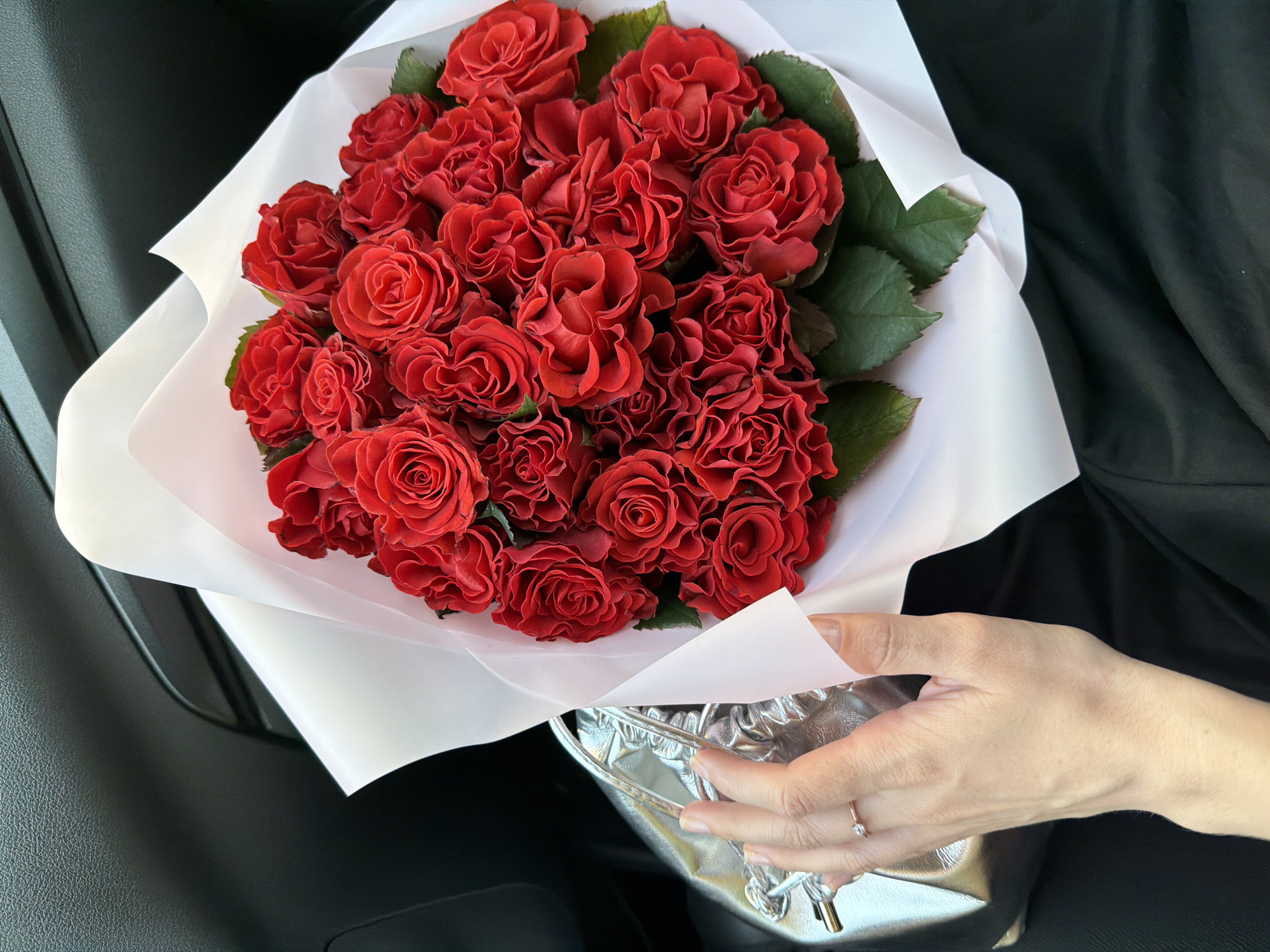 A high-angle, medium shot captures a bouquet of red roses wrapped in white paper, partially held by a person’s hand wearing a ring. The bouquet consists of numerous tightly packed roses with some green leaves visible. The hand is holding the bouquet above a reflective, metallic silver bag. The background includes parts of a black car interior. The location is not discernible.