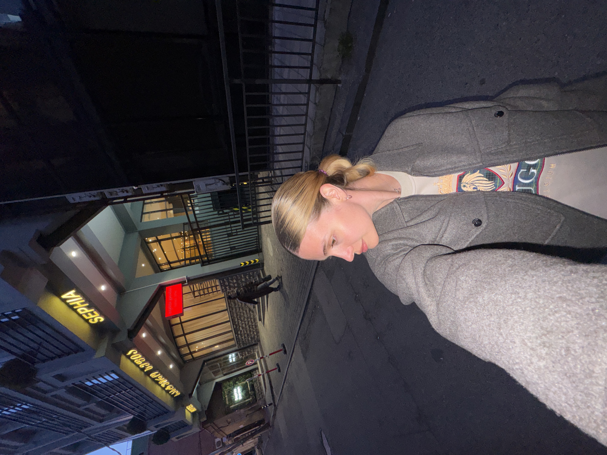 A person is taking a selfie on a street in Tbilisi, Georgia, at night. The person has blonde hair pulled back in a bun, and is wearing a gray coat over a light-colored top. The background includes a street, a building with "SEPHIA" in yellow letters, and a red sign that reads "Sephia Garden" in English and Georgian script. There is also a person walking in the background.