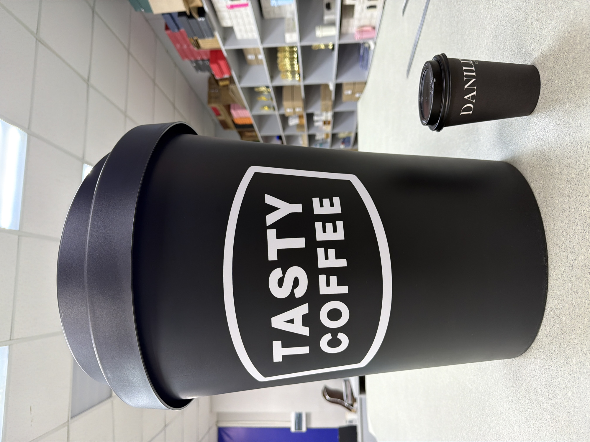 A rotated eye-level shot shows an extra-large, black coffee cup with a white logo that reads "TASTY COFFEE," next to a small, black coffee cup with a black lid, that reads "DANILI COFFEE" in white. The cups sit on a speckled white surface. In the blurry background, shelves stocked with boxes and small items can be seen. The location is Chaikovsky, Russia.