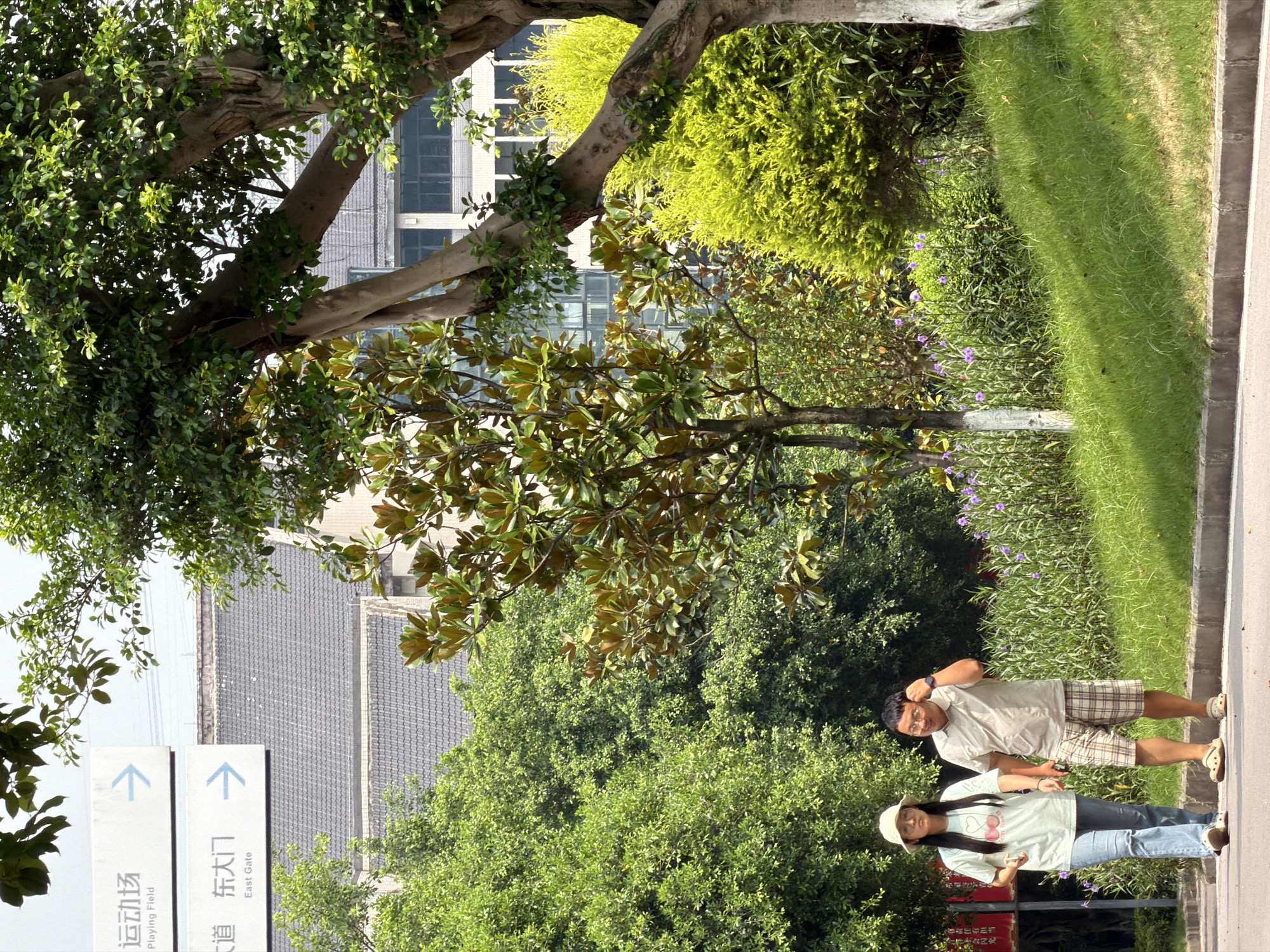 In the city of Jiangjin, China, a man and a woman are walking on a paved path next to a grassy slope. The man, wearing a light beige polo shirt and plaid shorts, is on the right, holding his right hand to his ear. The woman, on the left, is wearing a light green t-shirt with a cherry design and blue jeans, with a white bucket hat. They are walking away from the camera. Behind them, a dense green hedge with purple flowers is visible, interspersed with trees. In the background, a modern building with multiple windows and a dark tiled roof can be seen, partially obscured by the foliage. Two signs are mounted on poles to the left of the frame: one with Chinese characters and "Playing Field" written below, accompanied by a blue arrow pointing right, and another with Chinese characters and "East Gate" written below, also with a blue arrow pointing right. The overall environment appears to be a park or campus area.