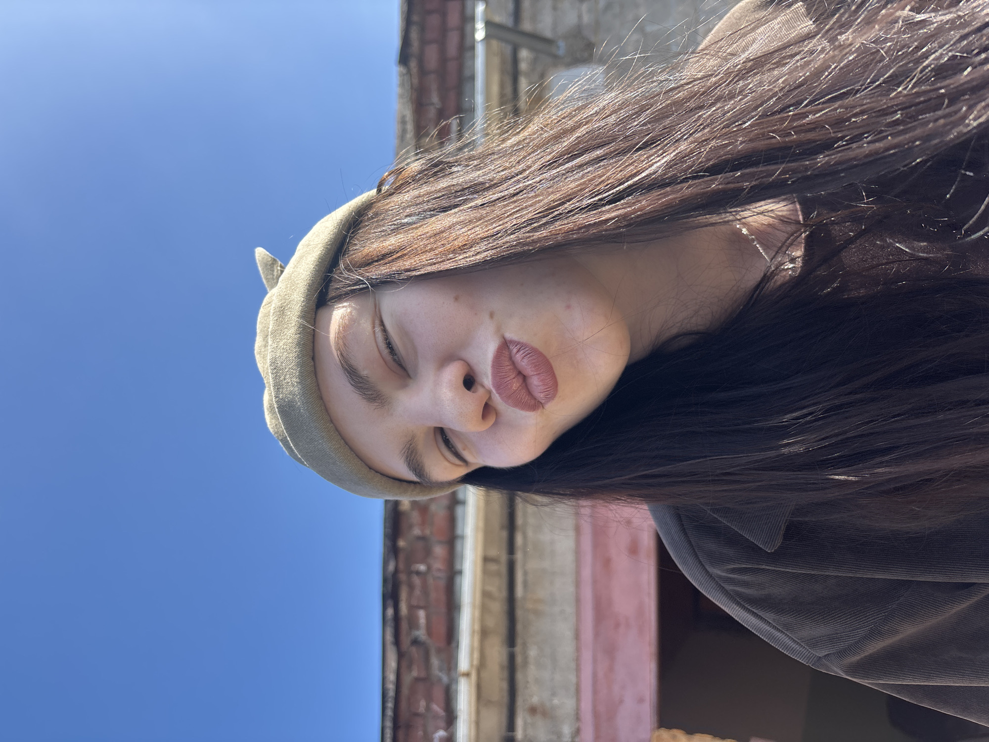 A young woman is centered in the frame, posing outdoors in Perm, Russia. She is wearing a corduroy jacket and a green cat-ear hat. The woman has long brown hair and pursed lips. The background features a blue sky and a brick and stucco building with pink and white panels.