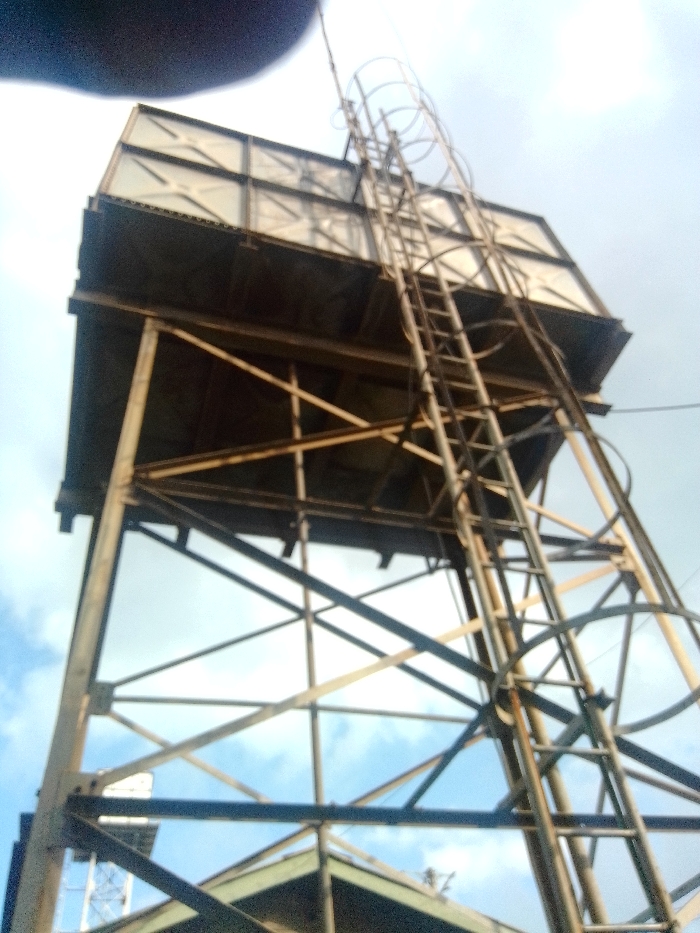 An elevated, square metal water tank is depicted, supported by a weathered metal structural framework. A vertical ladder, equipped with a safety cage consisting of circular metal hoops, extends from the ground level to the top of the tank. The metal components exhibit signs of weathering and potential surface corrosion. The sky above is partially cloudy with patches of blue visible. No individuals are present in the image. The specific location within Nairobi, Kenya, cannot be determined from the visual information provided.