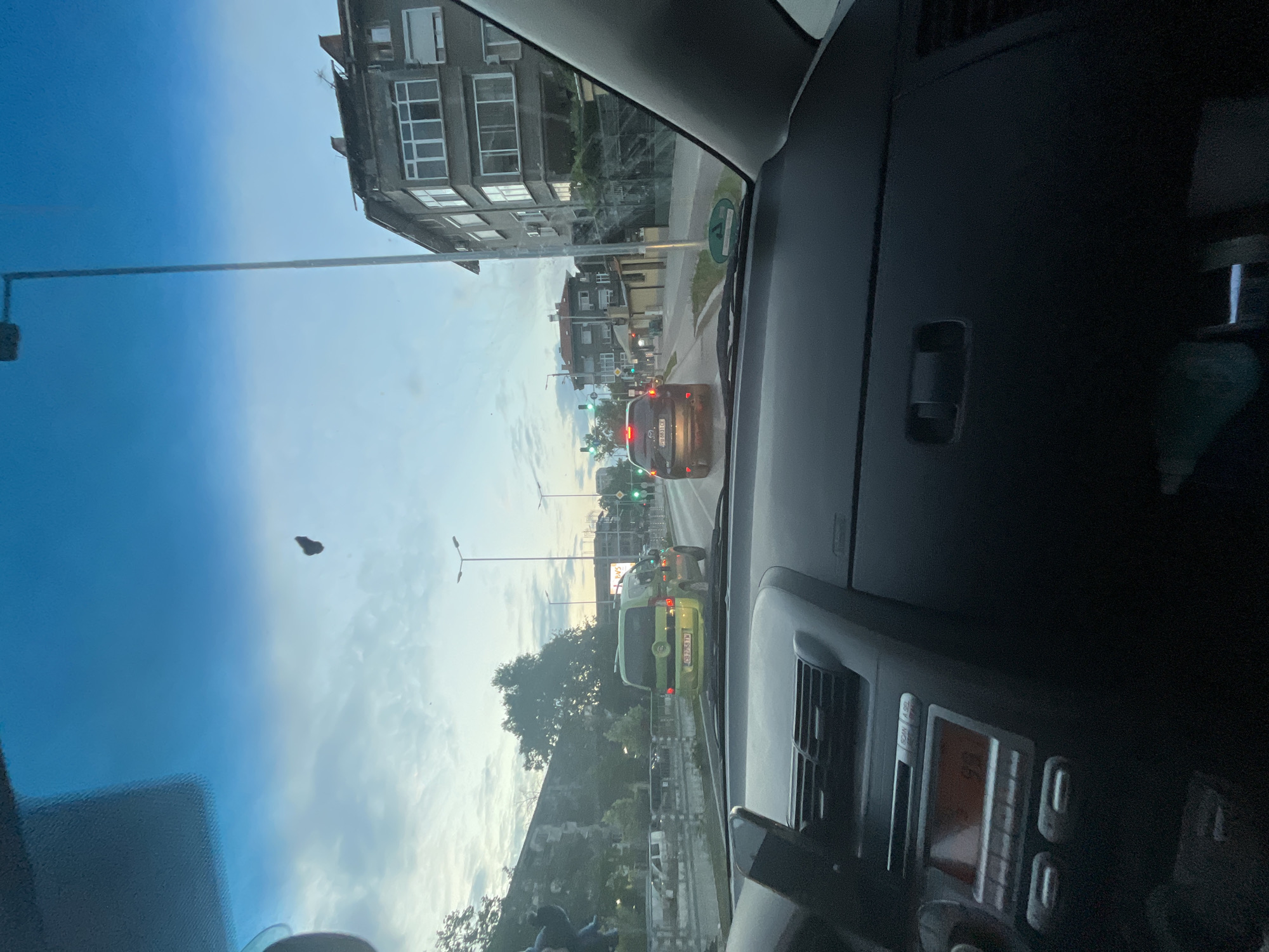 This is a first-person perspective media file taken from inside a car, looking out the windshield and side window. The scene is a city street with buildings, traffic lights, and other vehicles. The time of day appears to be dusk or early evening, with a dusky sky and a mix of natural and artificial light.
The subjects are primarily the urban environment and vehicles. The immediate foreground shows the car's interior, including a dashboard with a radio display. Outside the car, there are several vehicles visible on the road. A dark-colored car is in front, with its brake lights illuminated. Further ahead, a light green van is stopped at a traffic light. Buildings line the street, with apartment-style structures visible. A street lamp is also present. The sky shows a gradient of blue and gray, with some clouds. A dark, amorphous object is visible in the sky, possibly a bird or debris.
The activity taking place is likely commuting or driving through a city. The presence of traffic lights and multiple cars suggests a normal urban traffic flow. The overall mood is neutral, reflecting a typical drive.
Notable details include the car's radio display, which shows some text that is partially legible as "10:35" and possibly other symbols. The license plate on the green van is visible and reads "5738 AA". A circular green sticker with the letter 'A' is on the windshield of a vehicle in the distance, possibly indicating a parking permit or inspection sticker. The location context provided is Plovdiv, Bulgaria.