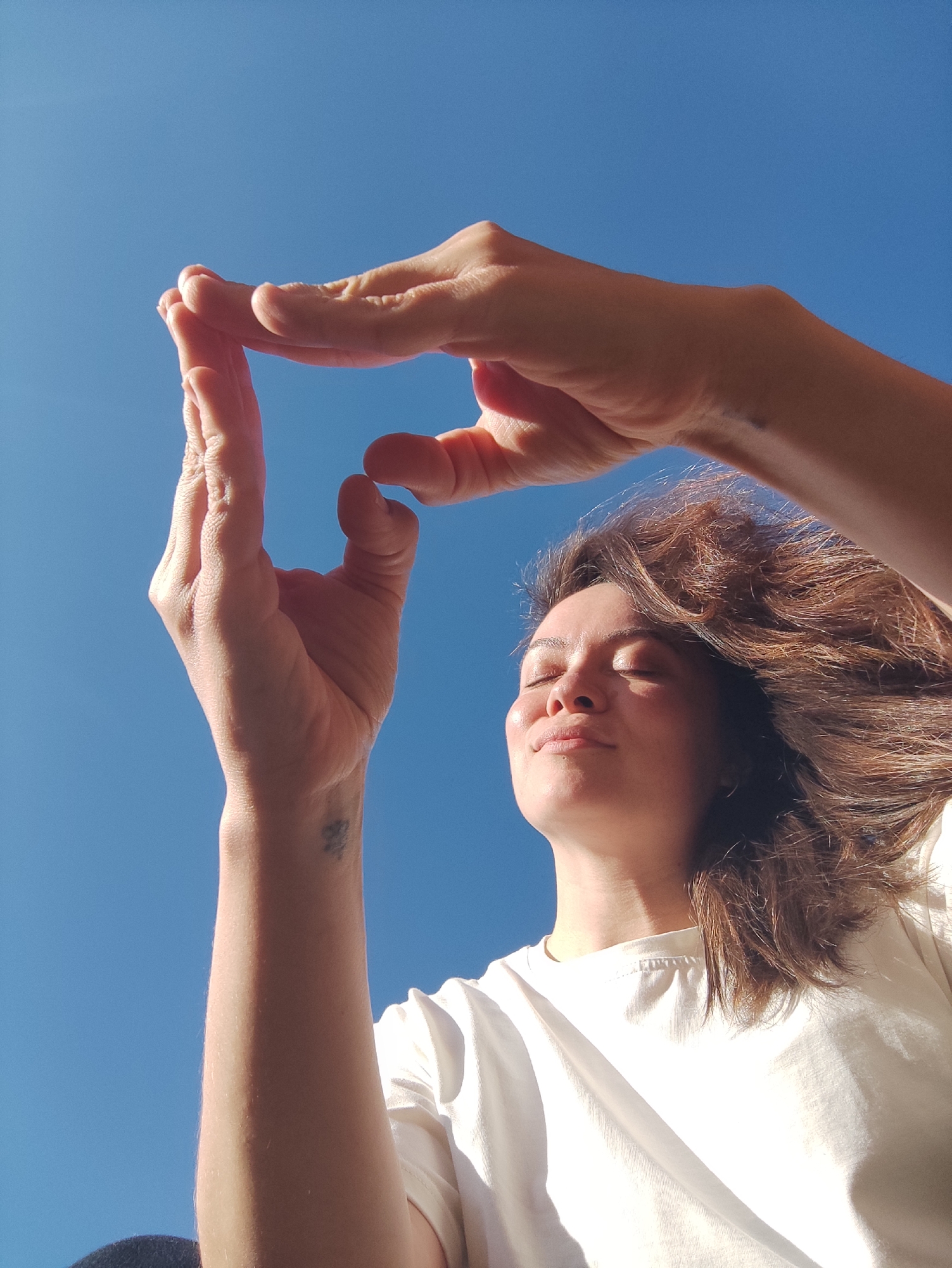 A low-angle portrait captures a young woman outdoors in Фоки, Russia, against a vibrant, cloudless blue sky. She is seen from the chest up, with her head tilted slightly back and her eyes closed, suggesting she is basking in bright sunlight. A serene and content expression rests on her face, with a hint of a smile. Her medium brown, wavy hair frames her face, appearing slightly wind-swept. She wears a plain, light-colored (likely white or off-white) t-shirt.
Her hands are raised above her head, with fingers curled to form a rectangular frame against the clear sky, as if framing a view or playfully engaging with the light. The strong sunlight casts bright highlights on her skin and hair. A small, dark, and faded tattoo, which appears to be an abstract design or stylized symbol, is visible on the inner wrist of her left arm. The scene conveys a sense of calm and enjoyment of the sunny weather, likely midday or early afternoon given the intensity of the light.