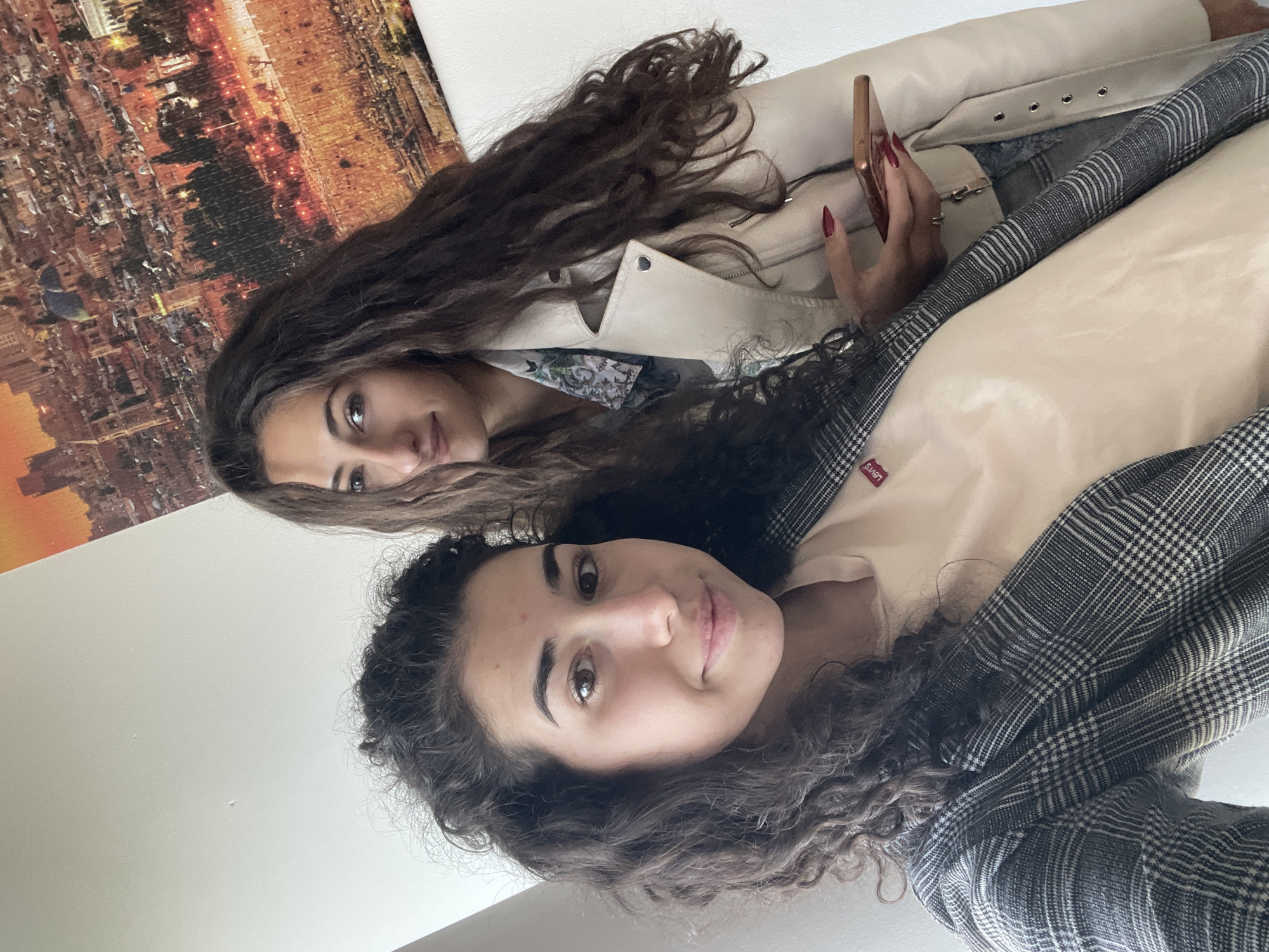 Two young women are posing closely together. The first woman, positioned at the top, has long, wavy dark hair and is wearing a light-colored jacket. She is holding a smartphone in her hand. The second woman, situated below, has curly dark hair and is wearing a plaid jacket over a light-colored top. The background features a blurred image of what appears to be a city skyline at night. The location cannot be determined to be in Sofia, Bulgaria from visual information.