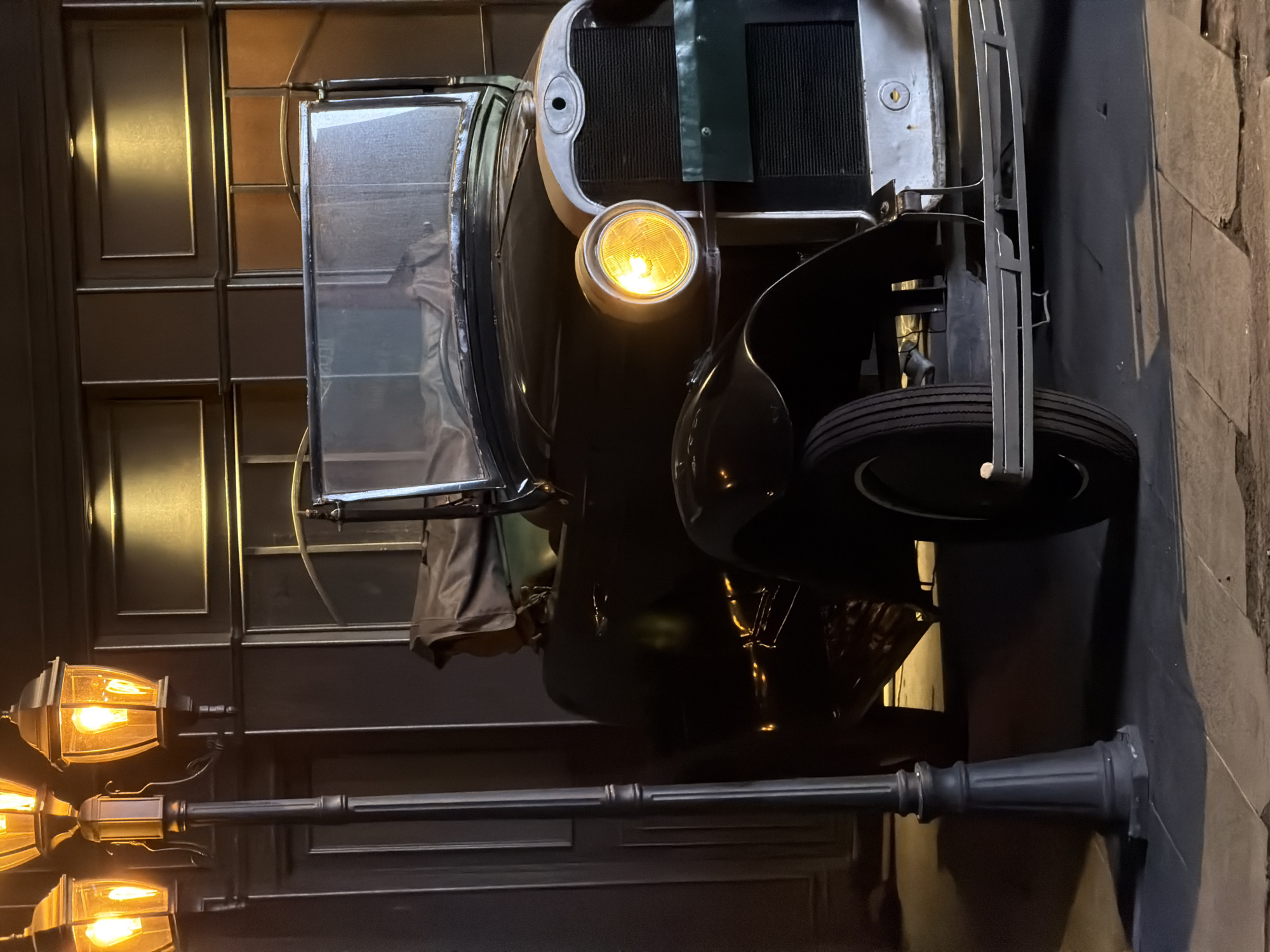 The image displays a vintage dark-colored automobile, possibly an antique car, parked next to a building facade under dim, atmospheric lighting, suggesting nighttime or an indoor themed environment. The car, seen from its front-left side, features a prominent, glowing yellow round headlight on the driver's side. It has a dark radiator grille encased in a silver-colored shell, and a portion of a spare tire is visible at the front passenger side. A folded soft top or cover is also discernible behind the windshield.
In the left foreground, a classic black street lamp with ornate detailing stands, holding three antique-style light fixtures, each emitting a warm, amber glow. The backdrop consists of a dark building wall with embossed or paneled sections, reflecting some of the ambient light. The overall scene conveys a nostalgic and potentially mysterious atmosphere. No individuals are present, and the car appears stationary, indicating no specific activity is taking place. There is no visible text within the image. The provided location context is North Kuta, Indonesia.