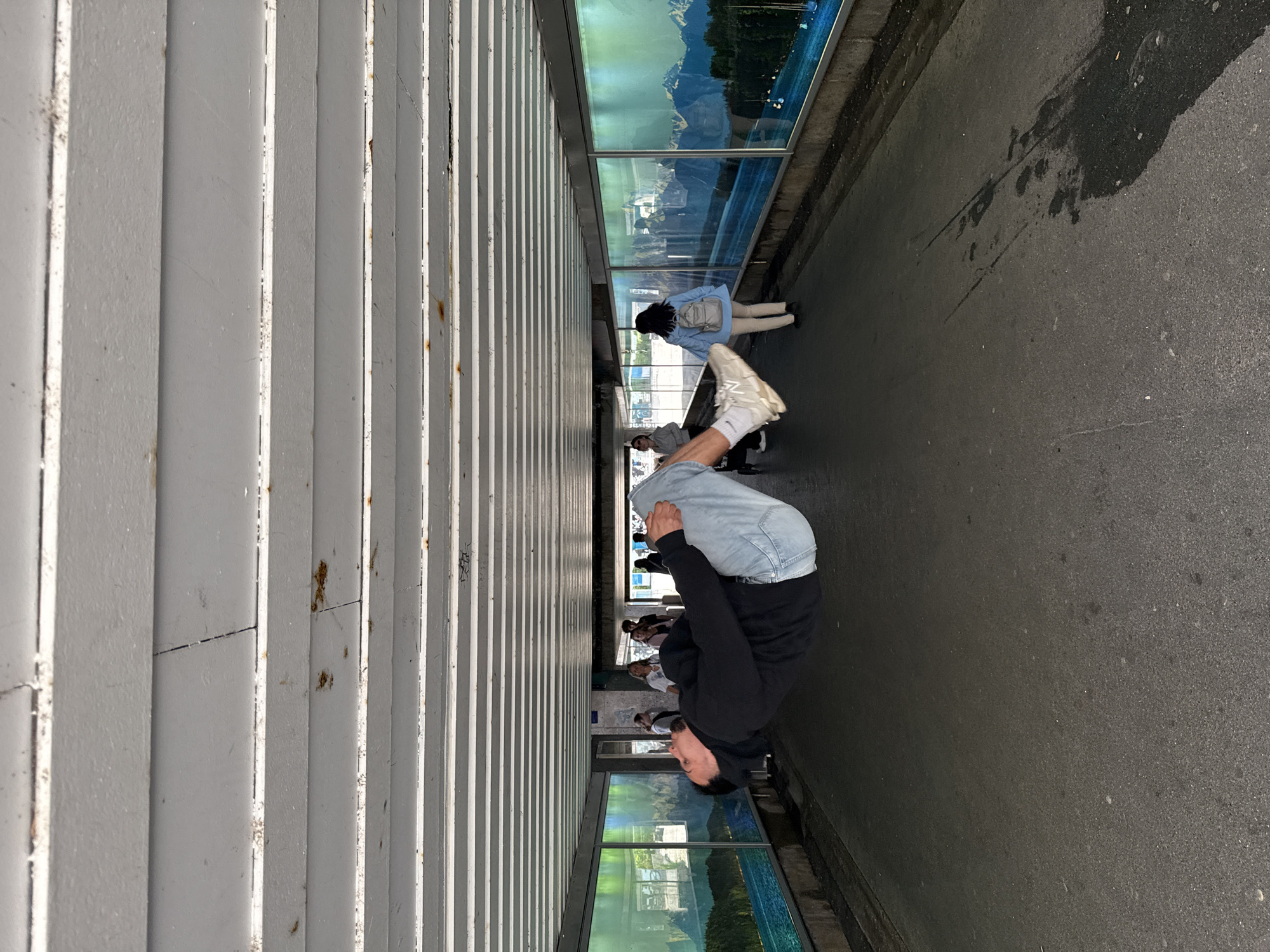 A man is captured mid-air, appearing to levitate in a covered walkway or underpass. He is wearing a black hoodie and light blue jeans, with his body curled inwards. His left leg is bent at the knee, and his right leg is extended. He is looking upwards and to his left.
The walkway has a white, ribbed ceiling and a grey, asphalt floor. On the walls of the walkway, large panoramic images are displayed, depicting a scenic landscape with mountains and a body of water, possibly a lake or fjord, under a sky with aurora borealis.
Several other people are visible in the background, walking in both directions. Some are blurred due to motion or distance. The overall lighting suggests it is daytime, with ambient light filtering into the walkway.
The location is identified as the Starnberg S-Bahn station underpass in Starnberg, Germany. The S-Bahn station is a public transport hub, and the underpass serves as a pedestrian connection. The presence of other commuters and the architecture of the walkway are consistent with such a location.