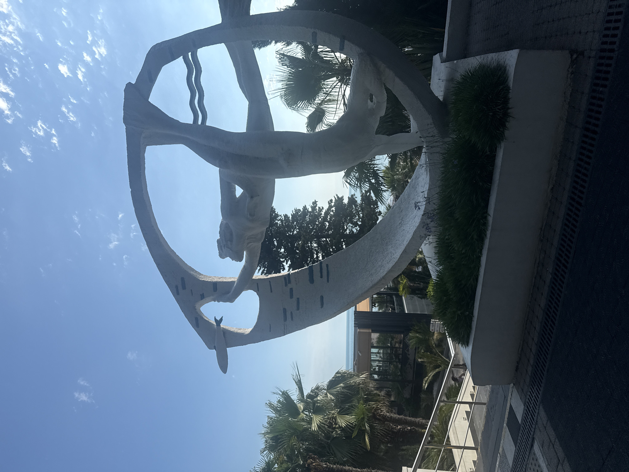 An outdoor sculpture depicting a human figure appears to be diving or swimming through a large, circular, abstract form. The sculpture is white and textured, possibly made of concrete or stone. It is positioned on a raised platform with lush green vegetation in front of it. Behind the sculpture, palm trees and other greenery are visible, with a building and the sea in the distance. The sky is a clear blue with scattered clouds. The sculpture is located in Sochi, Russia, specifically in front of the "Pearl of the Caucasus" hotel.