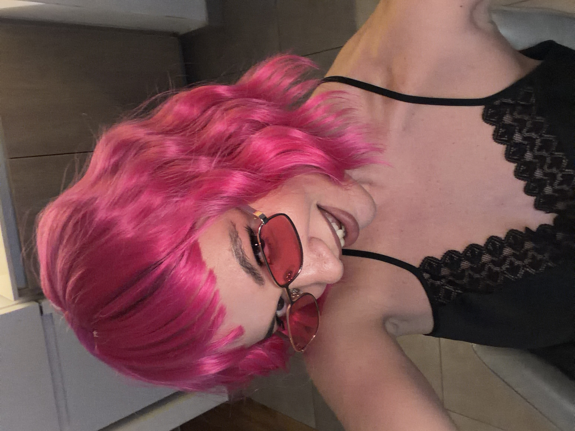 A woman with short, bright pink hair and red sunglasses is captured from a slightly angled, close-up perspective. She is wearing a black top with lace detailing along the neckline. The background is indistinct, suggesting an indoor setting.