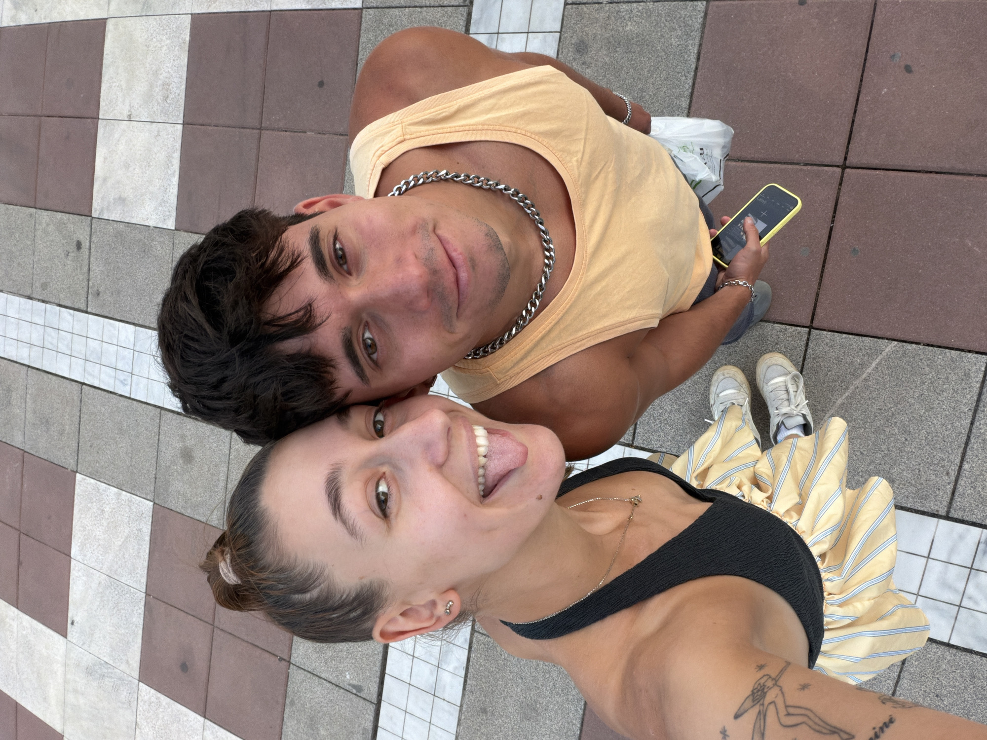 A high-angle, full-body shot shows a young couple taking a selfie outdoors. The woman, on the left, has her hair in a bun and is sticking her tongue out, wearing a black halter top and a yellow and white striped skirt. The man, on the right, is wearing a peach tank top and a thick silver chain necklace. He is holding a yellow-cased smartphone in his right hand and a white plastic bag in his left. The couple is standing on a checkered tile ground, which appears to be a public walkway or plaza. The background shows the continuation of the checkered pattern with a mix of reddish-brown, gray, and white tiles. The lighting suggests it is daytime. The scene is devoid of any specific identifying markers of Blagoevgrad, Bulgaria, beyond the implied context.