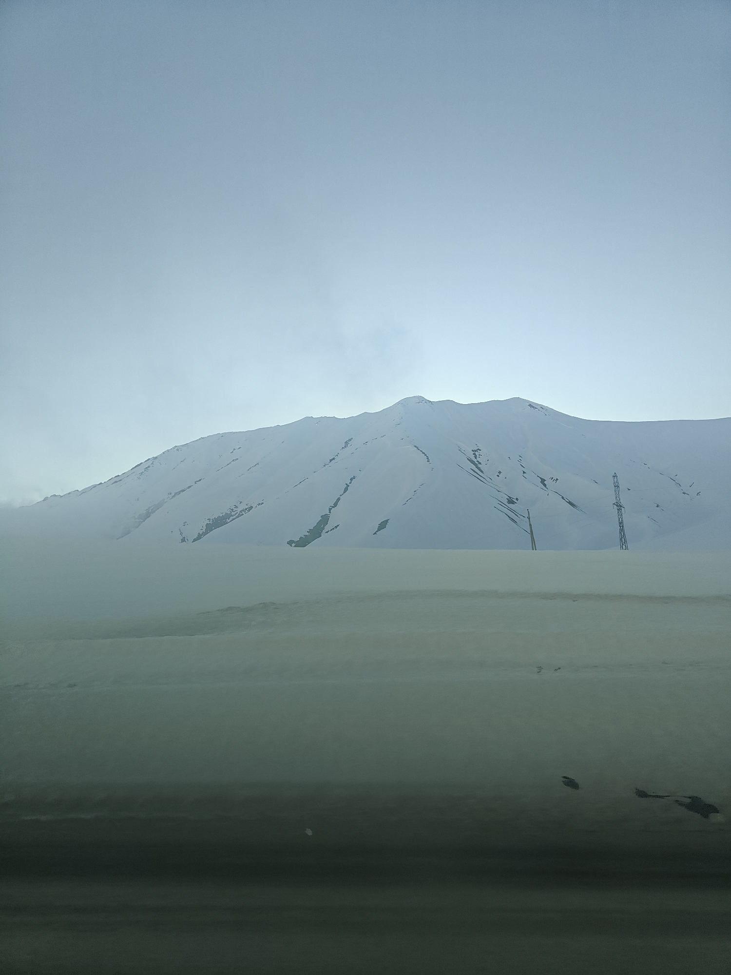 The image depicts a snow-covered landscape in what is likely the city of Ganisi, Georgia, based on the prompt. A large, snow-covered mountain dominates the background, partially obscured by haze or fog at its base. In the midground, a flat, snow-covered area is visible, punctuated by the presence of two power pylons/electrical transmission towers. The foreground features a dark, indeterminate surface, possibly a road or the interior of a vehicle, with indistinct dark shapes. The sky is overcast, contributing to the overall monochromatic and subdued tone of the scene. No people or animals are visible.