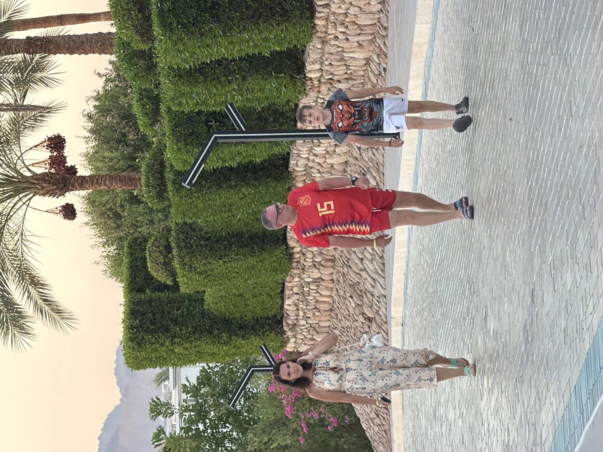 A medium shot captures three people walking on a paved pathway in an outdoor resort setting. The walkway, composed of interlocking gray pavers with blue and white trim, is lined by a low stone wall and manicured green hedges. Palm trees with dates hanging from them are visible in the background, as are distant mountains.
The individuals are walking from left to right. On the far left is a woman wearing a light-colored, patterned jumpsuit and light blue sandals. She is looking towards the camera and has her left hand near her face. Next to her, in the center, is a man in a red short-sleeved shirt with a Spanish flag motif and the number "15" on the front, paired with red shorts. He is also looking forward. To the right of the man is a young boy wearing a black t-shirt with a tiger graphic, white shorts, and dark sneakers. He is looking ahead. Two modern outdoor lamps with black frames and white accents are positioned behind the group, integrated into the hedge line. The lighting suggests it is either early morning or late afternoon. The scene appears to be in Sharm El Sheikh, Egypt, given the presence of palm trees, mountains, and resort landscaping.