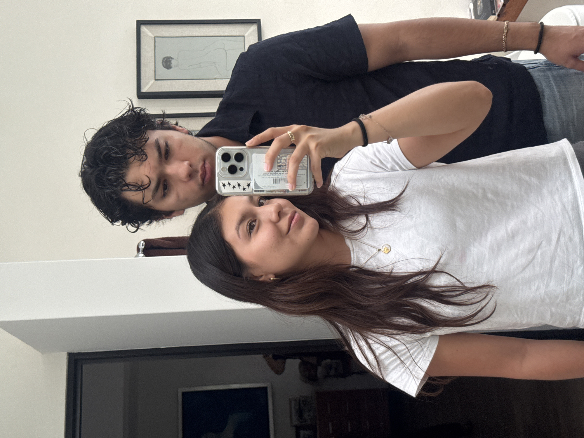 A selfie-style photograph shows two individuals, a male and a female, inside an interior space in Oaxaca City, Mexico. The female is holding a smartphone with a clear case adorned with black stars, taking the picture. She is wearing a white t-shirt and a necklace with a hexagonal pendant. Her dark brown hair falls across her shoulder. She is looking directly at the camera. The male is wearing a black shirt and is positioned behind her. He is also looking at the camera. A framed artwork hangs on the wall behind them, possibly a line drawing. The room appears to be part of a residence, with a doorway leading to another area visible in the bottom left. An additional picture hangs behind the woman and on a table. The lighting is even, with the image appearing to be taken indoors during the daytime. Both people are wearing bracelets. The man is wearing a gold one on his wrist and another is black. The woman is wearing two bracelets on her wrist as well.