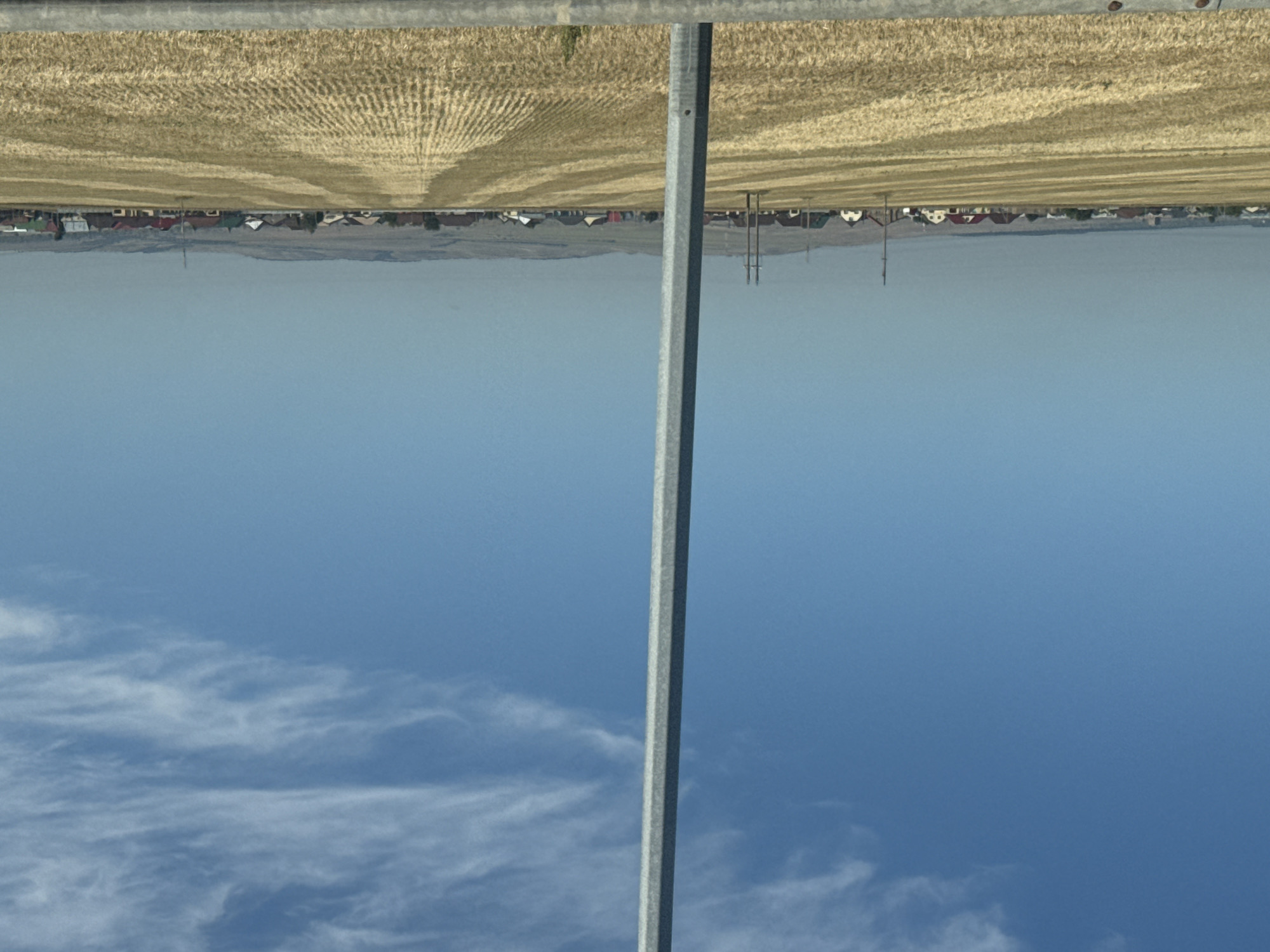 The image depicts a wide, rural landscape under a clear blue sky with wispy clouds. In the foreground, a tall, grey, octagonal pole stands vertically, dividing the frame. Beyond the pole, a vast field of harvested wheat or similar grain is visible, with distinct rows and stubble creating a textured pattern. In the middle ground, a long, horizontal line of houses with red, brown, and white roofs stretches across the horizon, indicating a settlement or village. Several utility poles and wires are interspersed within this village area. The background shows rolling hills under a hazy sky. The bottom edge of the image has a blurred, grey metallic barrier. This scene is representative of the agricultural outskirts of Sorochinsk, Russia, likely during harvest season. There are no people visible in the image.