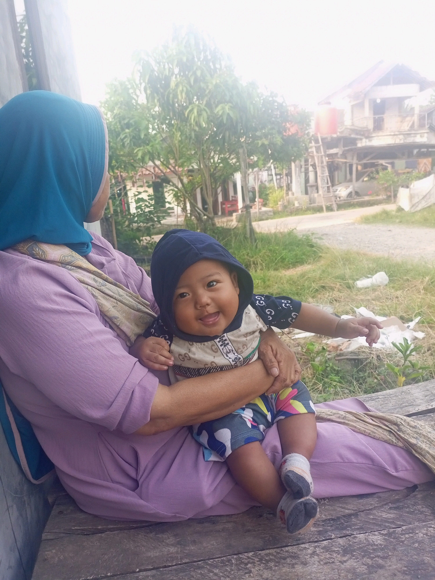 In Jawilan, Indonesia, a person wearing a blue hijab and a purple top sits on a wooden bench, holding a baby. The baby, wearing a hooded shirt and colorful shorts, smiles and reaches out a hand. The background shows a rural scene with trees, houses, and a dirt road.