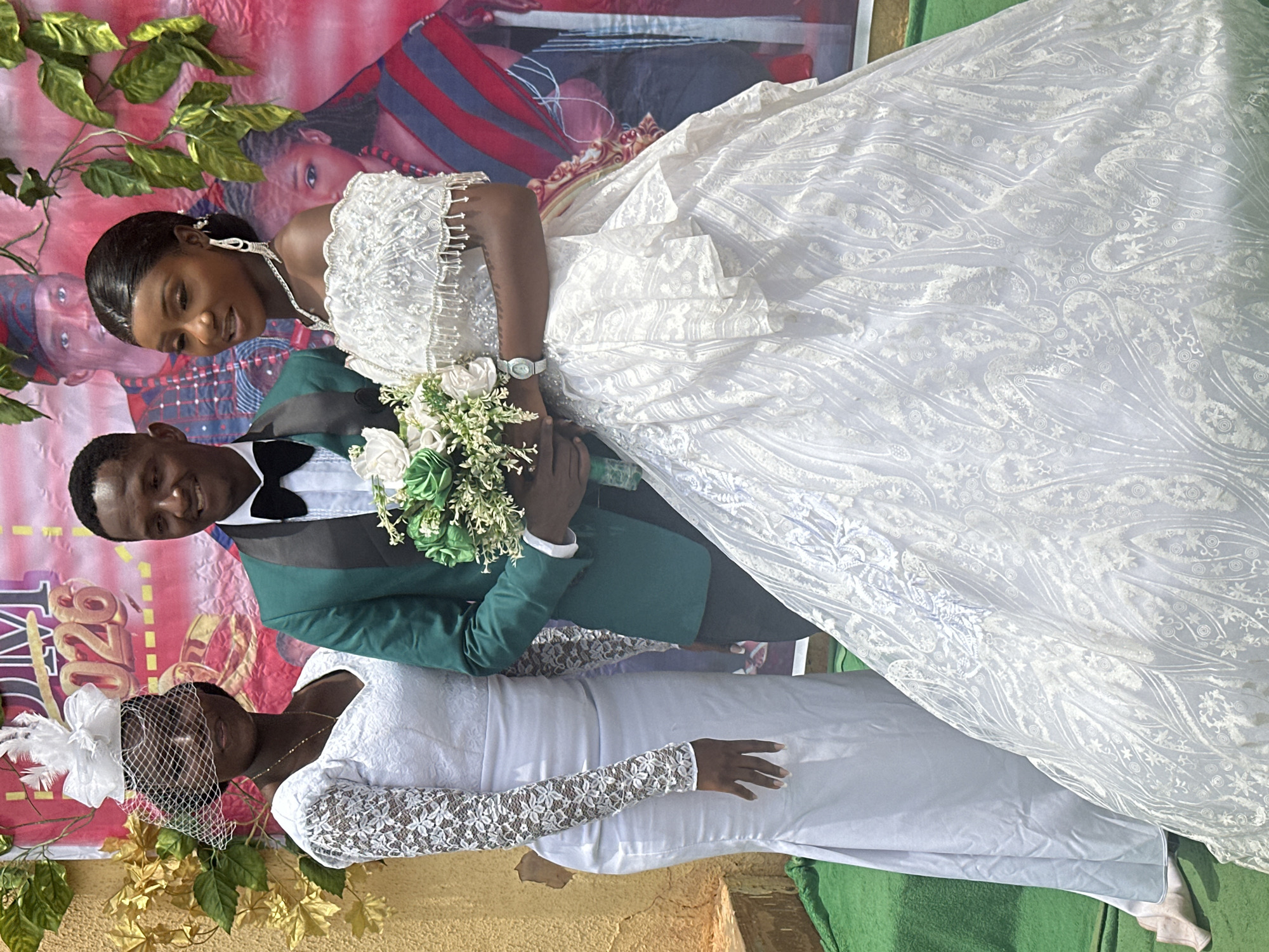 The image captures a wedding scene in what appears to be Unknown, Unknown. The focal point is a newly married couple. The bride, adorned in a white wedding gown with intricate lace patterns and beaded detailing, is holding a bouquet of white and green flowers. Beside her stands the groom, dressed in a green suit with a black bow tie. An older woman, presumably a relative, is also present, wearing a white dress with lace sleeves and a white fascinator.
Behind the group is a banner featuring images of people in ethnic attire, suggesting a celebration of cultural heritage. Green vines and decorative leaves frame the upper left corner of the image. The background appears to be an outdoor setting with a green fabric on the ground. The overall environment suggests a modest wedding celebration.