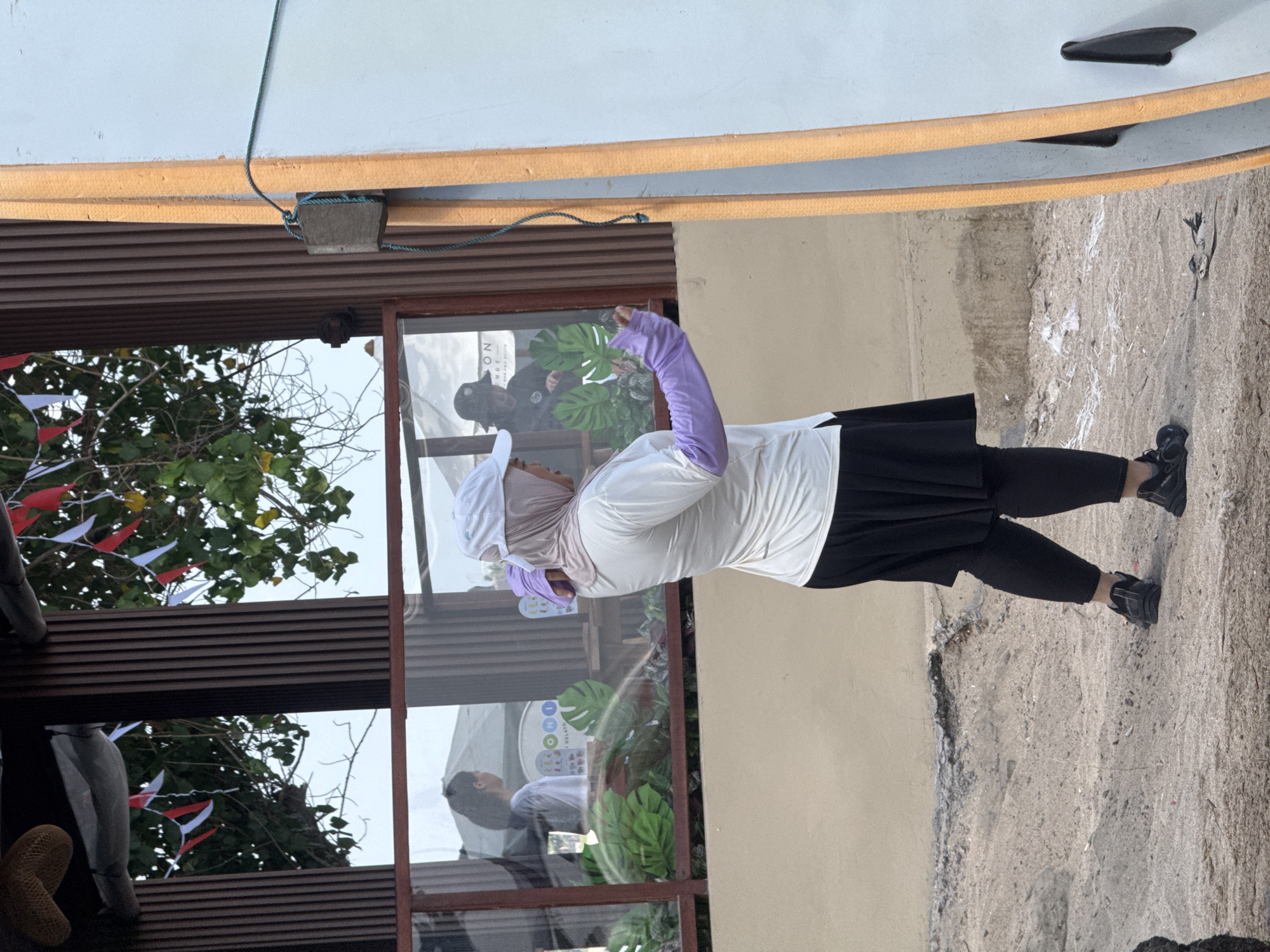 A woman wearing a white hijab, a white t-shirt, purple sleeves, and a black skirt stands in front of a building. She is holding a phone in her right hand, and her left hand is extended. The building appears to be a cafe or restaurant with large windows, through which other people are visible. The exterior wall of the building is beige. In the foreground, there is a sandy area with some debris. To the right of the woman is a light blue surfboard with an orange edge. The background shows trees and a sky. The scene is likely in Kuta, Indonesia, given the presence of surfboards and the architectural style.