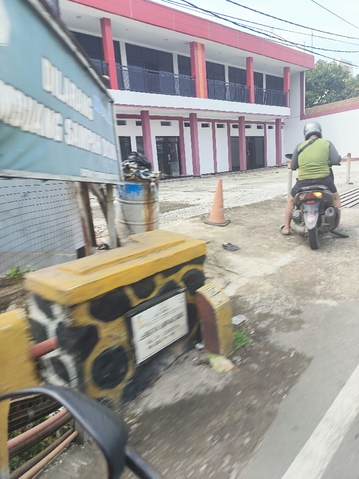 A man wearing a lime green shirt and a helmet rides a motorcycle in front of a building in Ngamprah, Indonesia. The building has red columns and a white facade. The man is riding towards the camera. There is a black trash can and a pile of garbage in the street. A traffic cone is on the ground near the curb. The ground is paved. There is a yellow and black painted concrete barrier to the left of the image. A sign is visible with the Indonesian word "DILARANG" on it. The view is from a car in the street. The coordinates are -6.854, 107.498.
