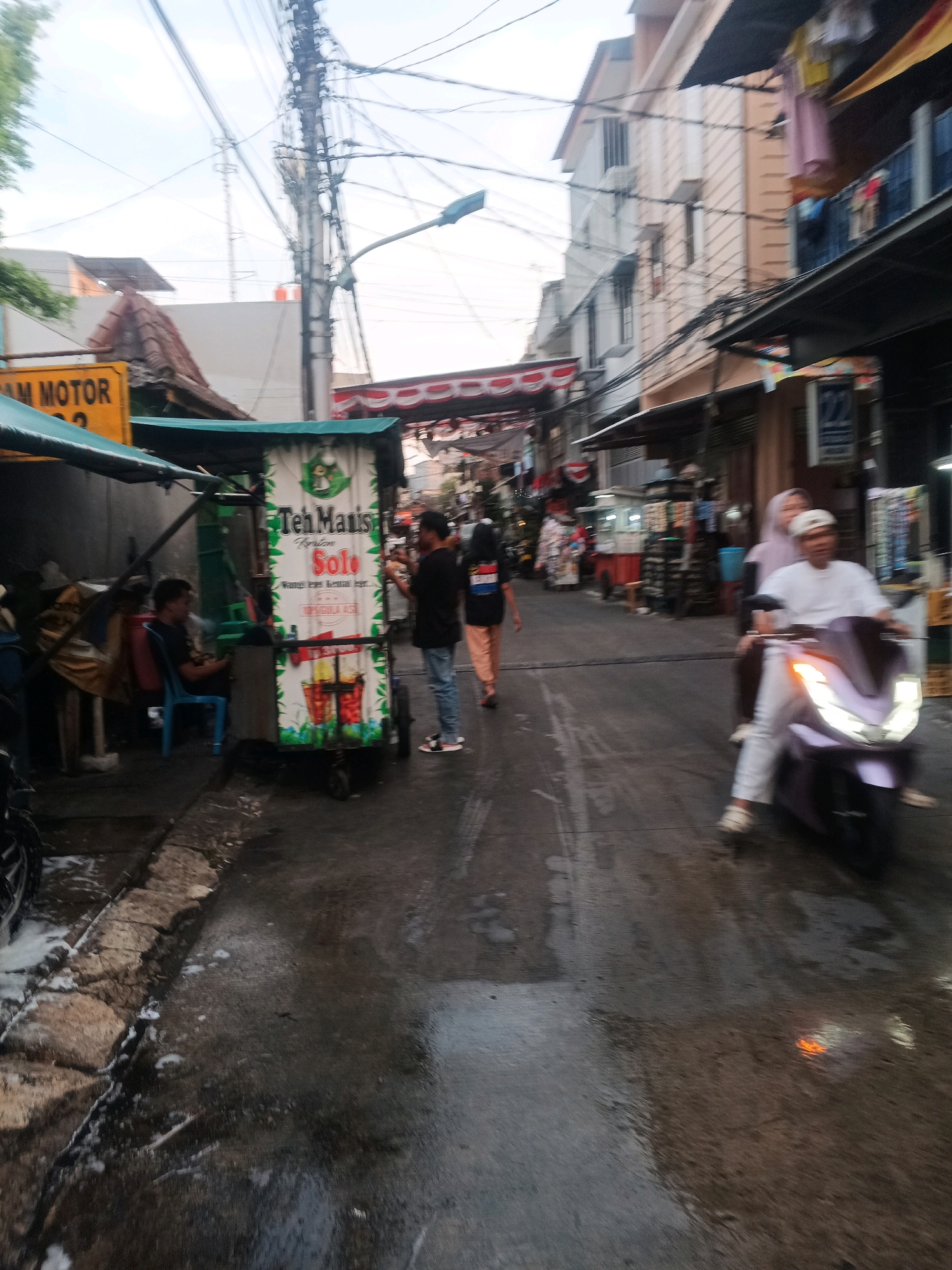 The image depicts a street scene in West Jakarta, Indonesia, likely a market or commercial area, with a focus on a street food cart. The street is wet and appears to have recently rained, with reflections on the asphalt. On the left, a yellow sign reading "JAM MOTOR" (motorcycle parking) is visible. A small food cart, branded "Teh Manis Solo," selling tea, is prominent in the center, with images of beverages on its side. A person is seated on a blue chair next to the cart. Two individuals, one in a black shirt and jeans and another in a blue shirt and jeans, are standing near the cart. Further down the street, a person on a light purple scooter, wearing white clothing and a white cap, is riding towards the camera, with a passenger behind them wearing a light pink headscarf. In the background, buildings with multiple stories are visible, some with awnings and signs. Laundry hangs from some of the buildings. The overall impression is of a bustling urban environment.
