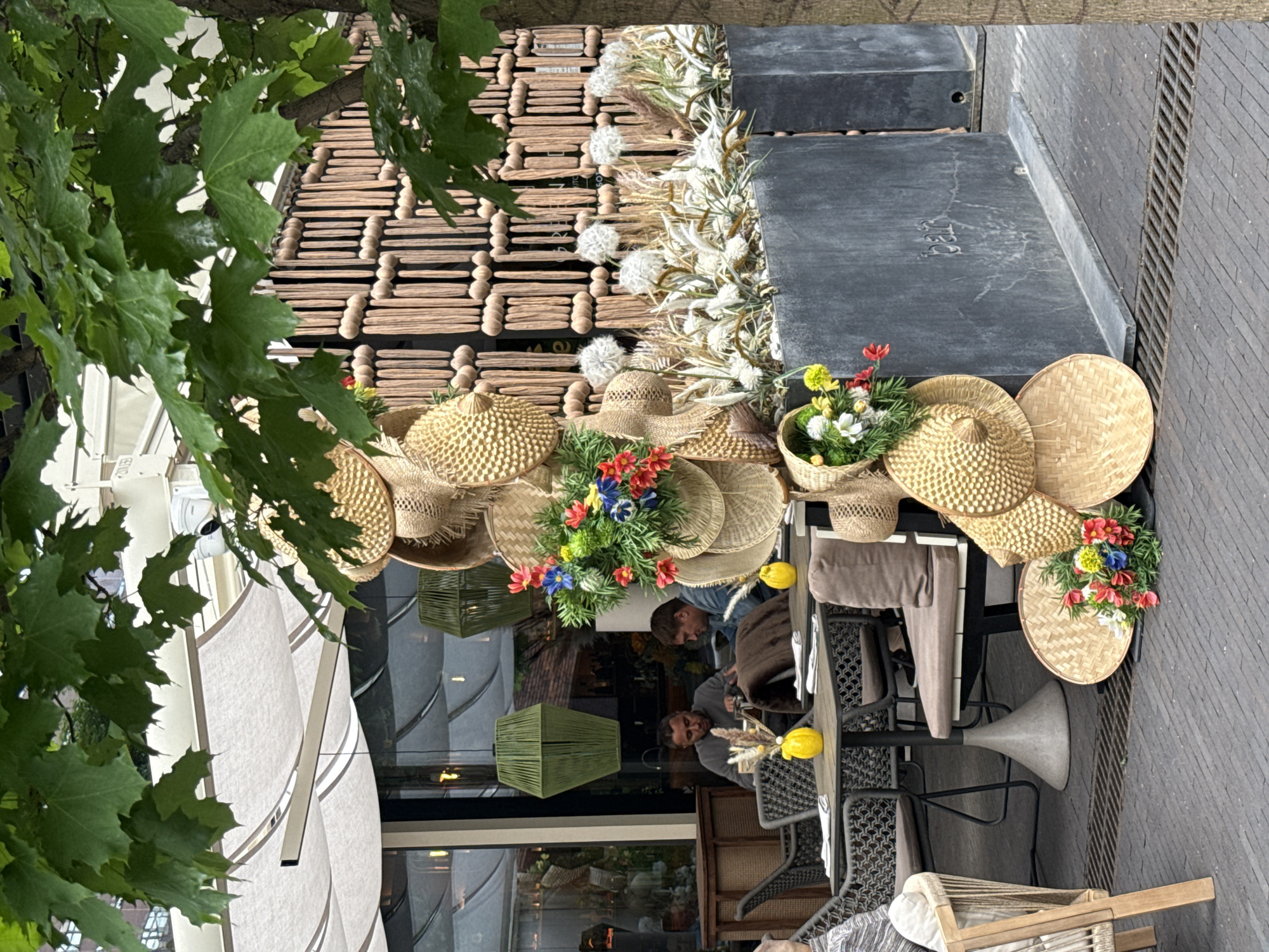 The provided image, rotated 90 degrees counter-clockwise, depicts an inviting outdoor cafe or restaurant patio scene, likely in an urban setting during daylight hours. Lush green tree leaves partially frame the top and left side of the frame, suggesting a tree-lined street.
The foreground features several outdoor tables and chairs, some with light-colored cushions and others in a wicker or rattan style, set on dark paved ground. A large, light-colored patio umbrella is partially visible on the far left. The cafe is richly decorated with numerous woven straw hats of various shapes and sizes, some displayed individually, others grouped together or adorning tables. Many of these hats, along with several baskets, are filled with colorful artificial flowers in shades of red, blue, yellow, and white. Yellow spherical decorations, possibly lights or fruit, are also placed on some tables. Along a dark grey ledge or planter wall, there's an arrangement of dried grasses and white, fluffy decorative elements.
Behind the outdoor seating area is the facade of a building, featuring a distinctive decorative wall made of wooden slats arranged in a grid pattern with rounded wooden pegs at intersections. Adjacent to this is a darker, possibly stone or concrete section. Below this, large glass windows reflect the interior of the establishment. Through these reflections, two men are visible seated at a table indoors: one man, with a beard, looks towards the right, while another in a denim jacket looks down. On one of the dark grey concrete blocks, faint, unclear embossed numbers, possibly "123," are visible.
The overall atmosphere is calm and pleasant, characteristic of an inviting outdoor dining experience. The time of day appears to be during daylight hours, but without direct sunlight, suggesting an overcast day or a shaded location.
