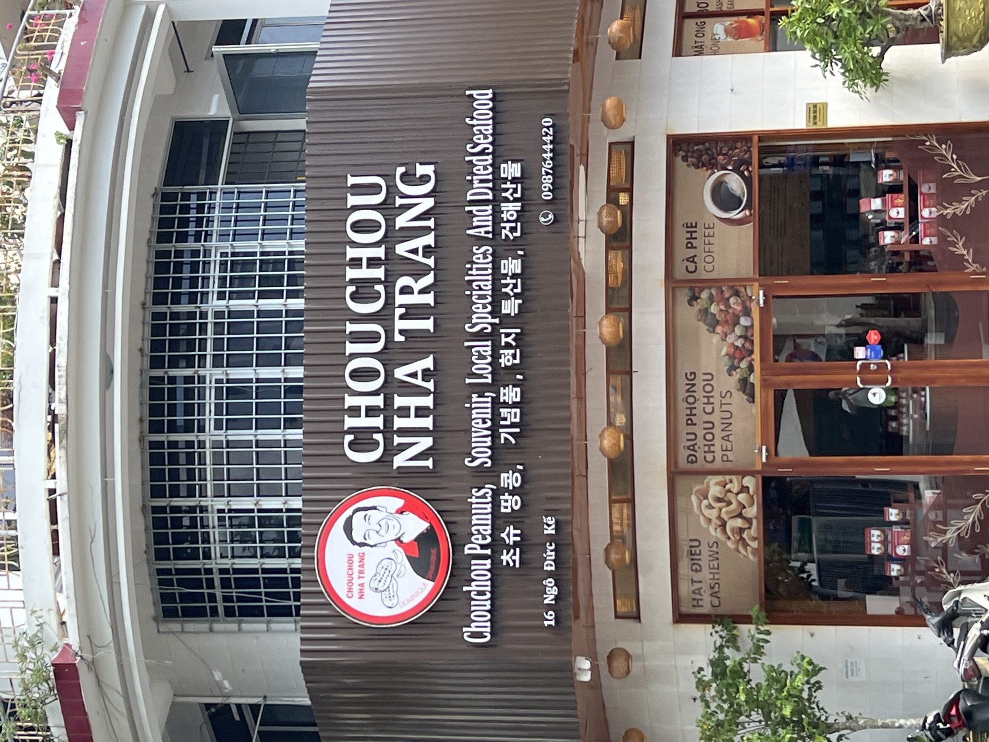 A street-level exterior shot captures the facade of a shop named "CHOUCHOU NHA TRANG" in Nha Trang, Vietnam. The shop's name is prominently displayed in large white lettering on a dark brown, corrugated metal background. Above this, a circular logo features a cartoon drawing of a smiling man with peanuts, and the text "CHOUCHOU NHA TRANG" and "DOMINIQUE SAUMONE".
Below the main signage, smaller text reads "Chouchou Peanuts, Souvenir, Local Specialties And Dried Seafood" in English, followed by Korean text and a phone number, "0987644420". The address "16 Ngô Đức Kế" is also visible.
The shop's windows display various products, including bags of cashews with the text "HAT DIEU CASHEWS", bags of peanuts labeled "ĐẬU PHÔNG CHOU CHOU PEANUTS", and a display of coffee with "CÀ PHÊ COFFEE". Another window shows products labeled "MẬT ONG BƠ HONEY" and what appears to be dried seafood. Inside the shop, displays of packaged goods are visible through the windows.
The upper part of the building features a rounded architectural element with metal grating and several windows. Decorative woven light fixtures hang from the overhang above the shop windows. A small tree is visible to the right of the shop, and a motorcycle is parked on the sidewalk to the left. The overall scene suggests a commercial establishment selling local Vietnamese products.
