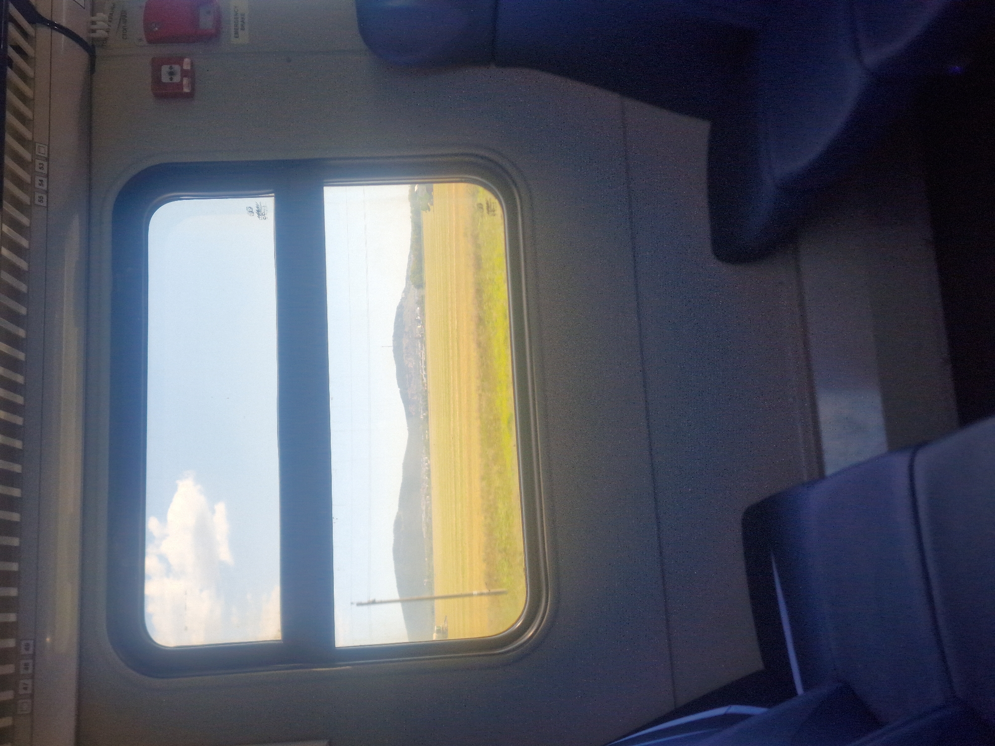 The image depicts the interior of a train carriage looking out a window. The window reveals a rural landscape with a vast, yellow field under a cloudy blue sky. In the distance, rolling hills with some buildings are visible. Inside the carriage, two dark blue seats are partially visible in the foreground, one on the left and one on the right, suggesting the perspective is from a seated passenger. The wall of the carriage is a neutral gray, with some emergency equipment, including a red fire alarm and an "EMERGENCY BRAKE" label, mounted on the right side. The ceiling has a ribbed pattern with small numbered labels. No people are clearly visible within the train carriage. The overall impression is of a peaceful journey through the countryside.